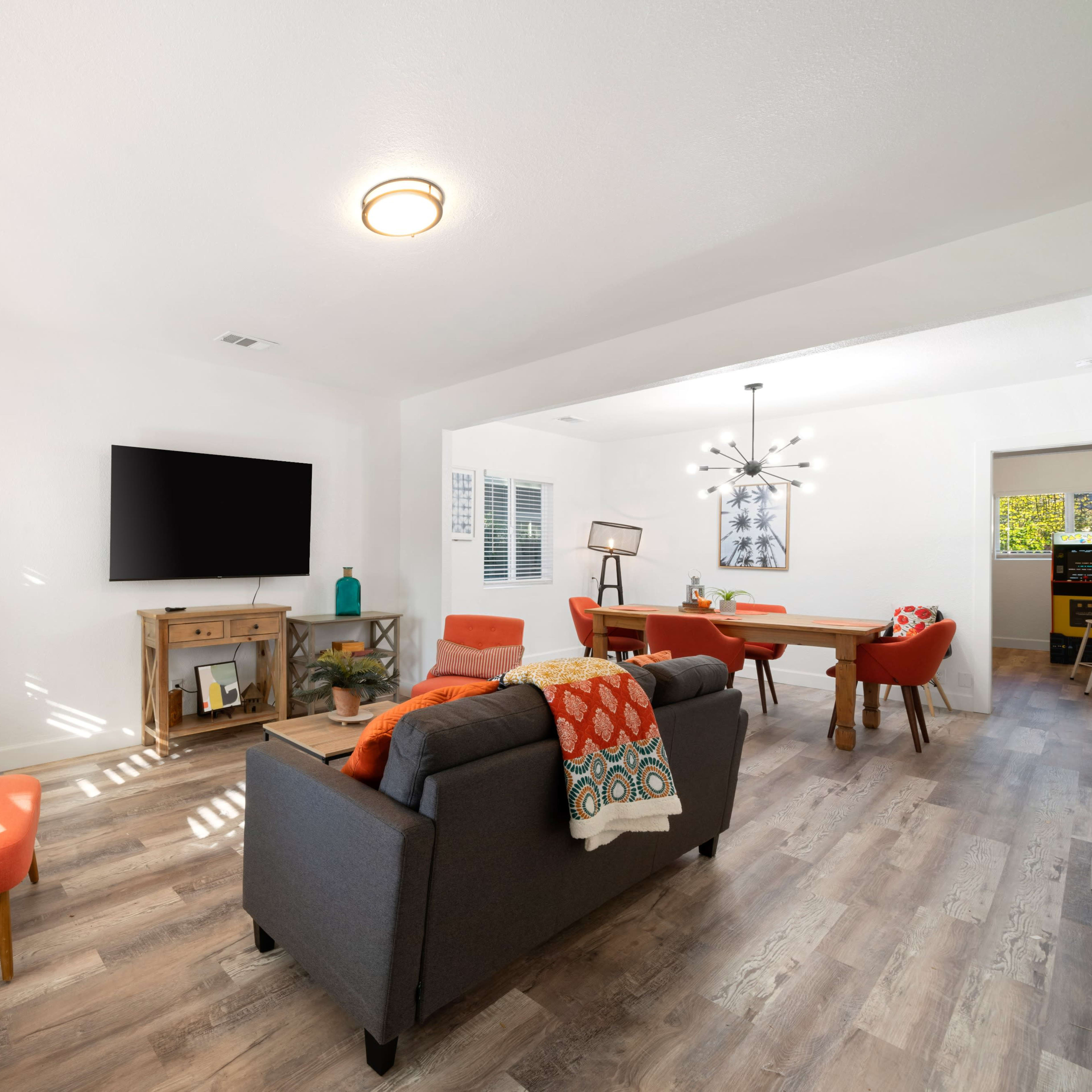 The living room features a gray sofa and two orange chairs, a wooden coffee table, a wall-mounted television, and a dining area with a light fixture.