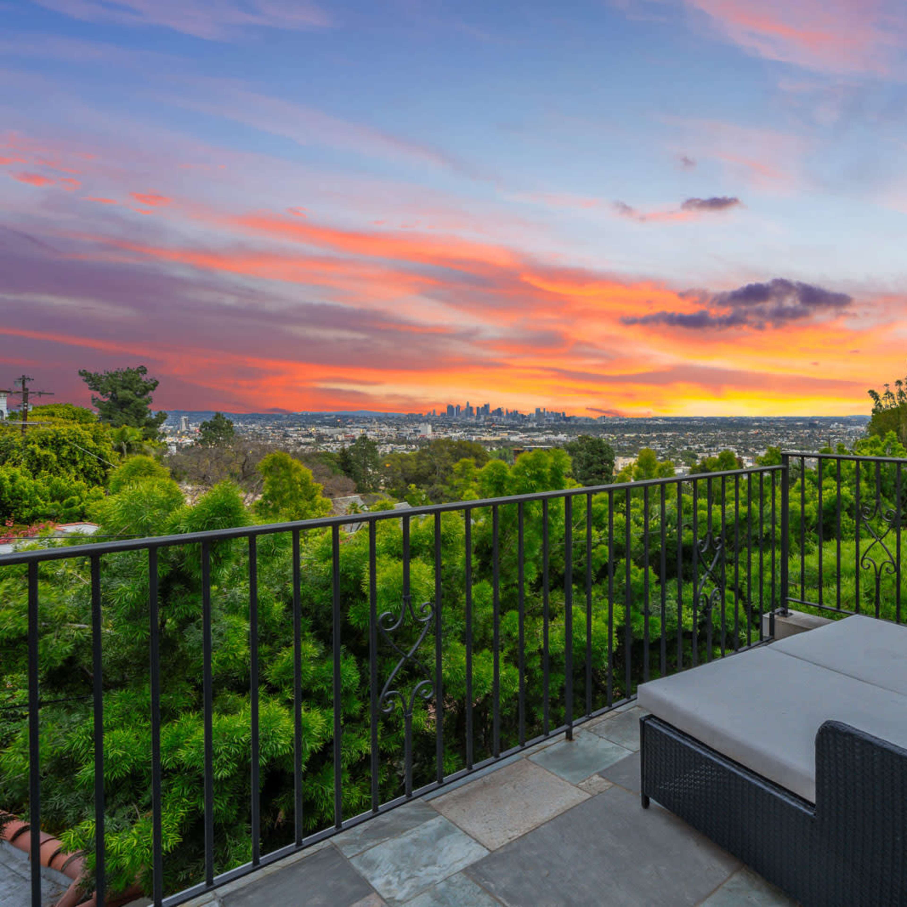 A balcony overlooking a city skyline at sunset, framed by lush greenery.