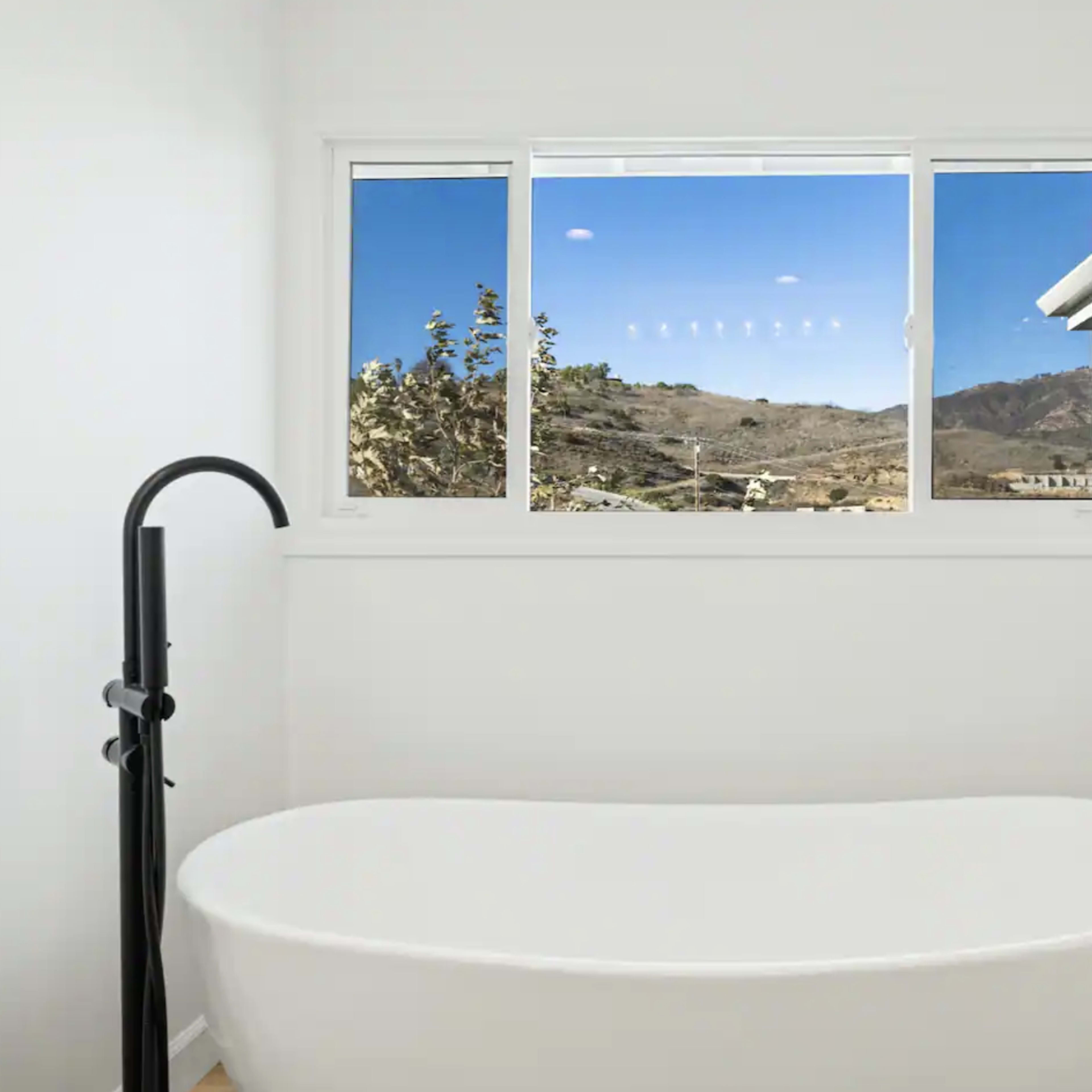 A modern bathroom features a freestanding white bathtub in front of a large window showcasing a landscape with mountains.
