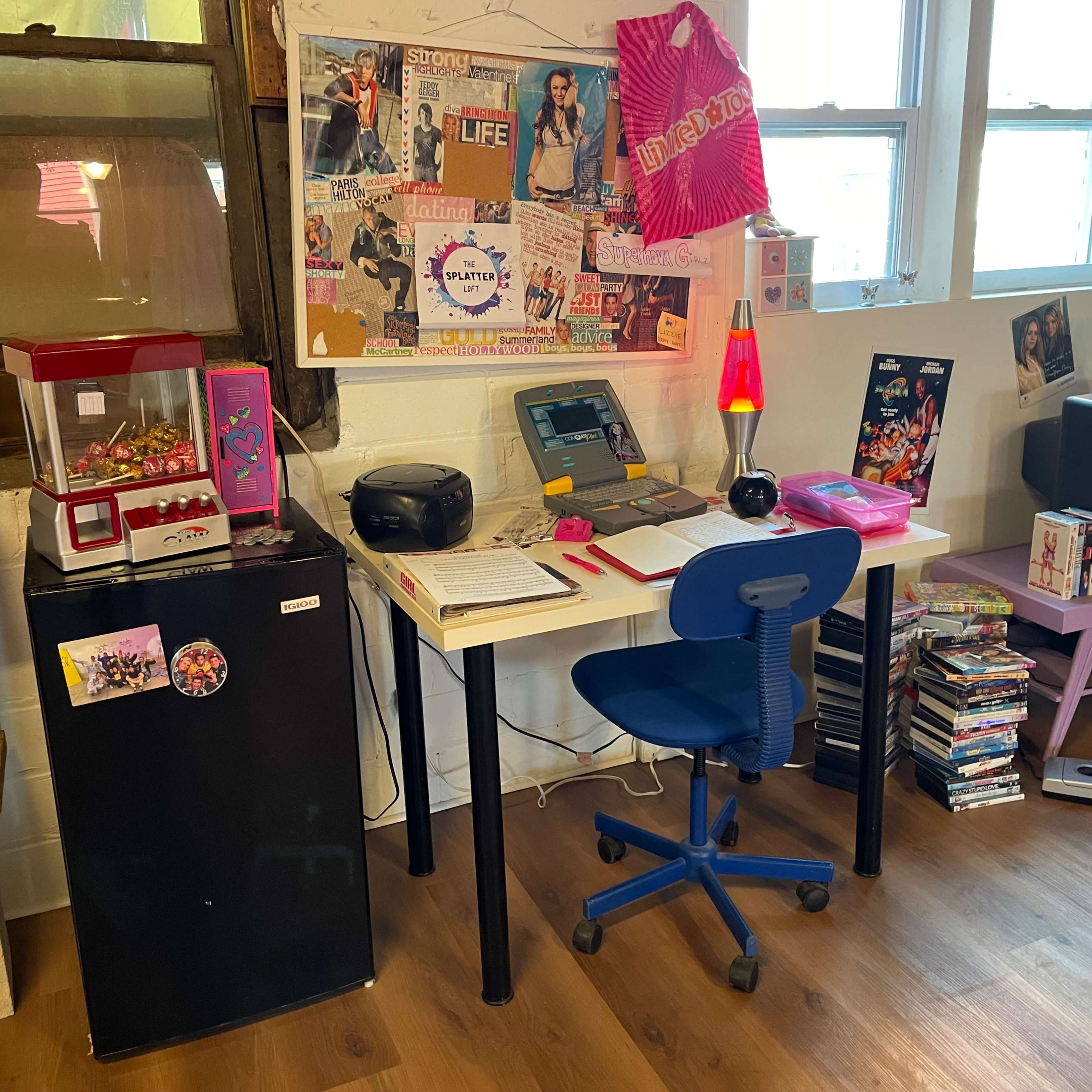 A small workspace with a desk, a blue chair, assorted vintage decorations, a television, and a collection of DVDs, alongside a mini fridge and a candy dispenser.
