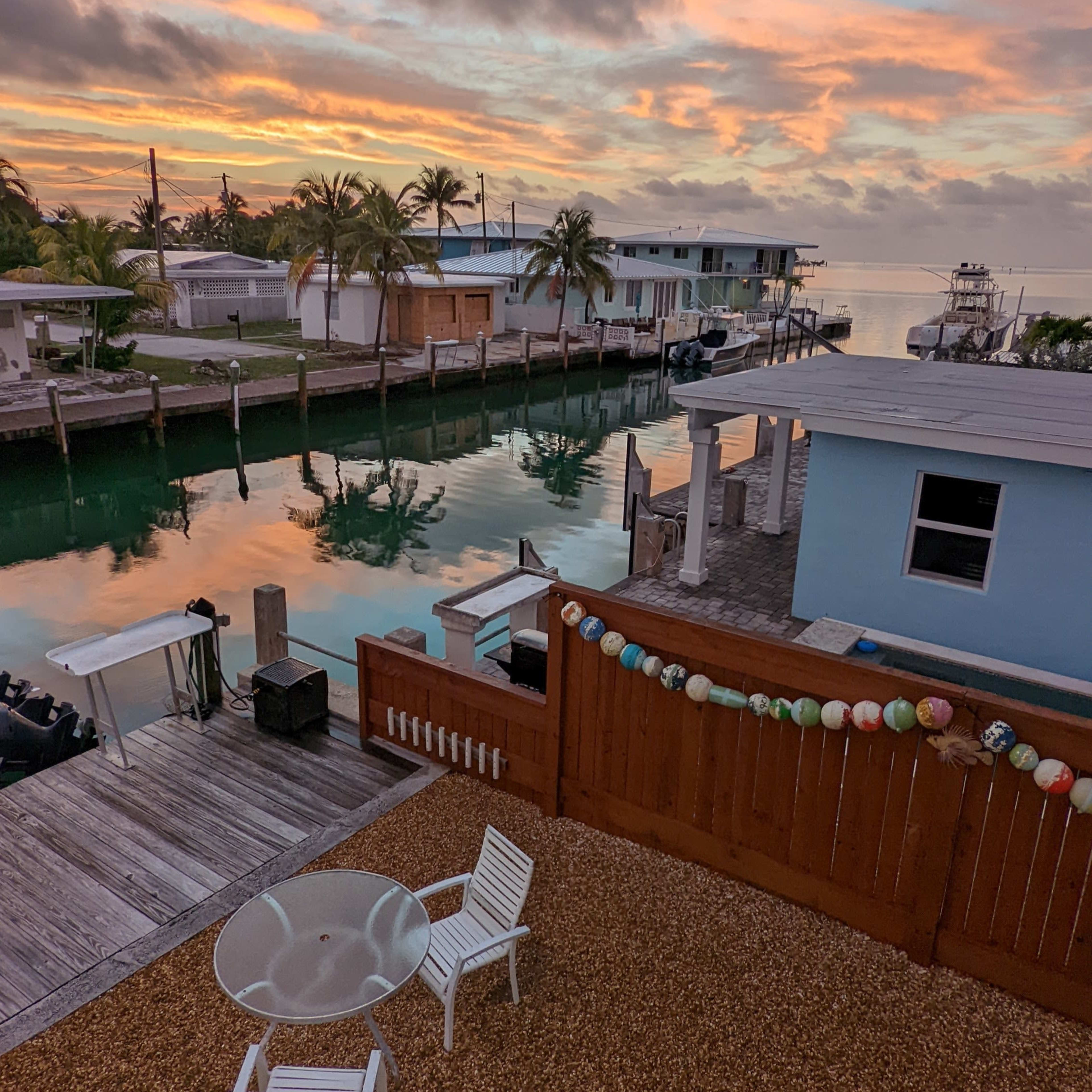 A waterfront scene at sunrise with a table and chairs in the foreground, surrounded by a gravel area and palm trees along the canal.