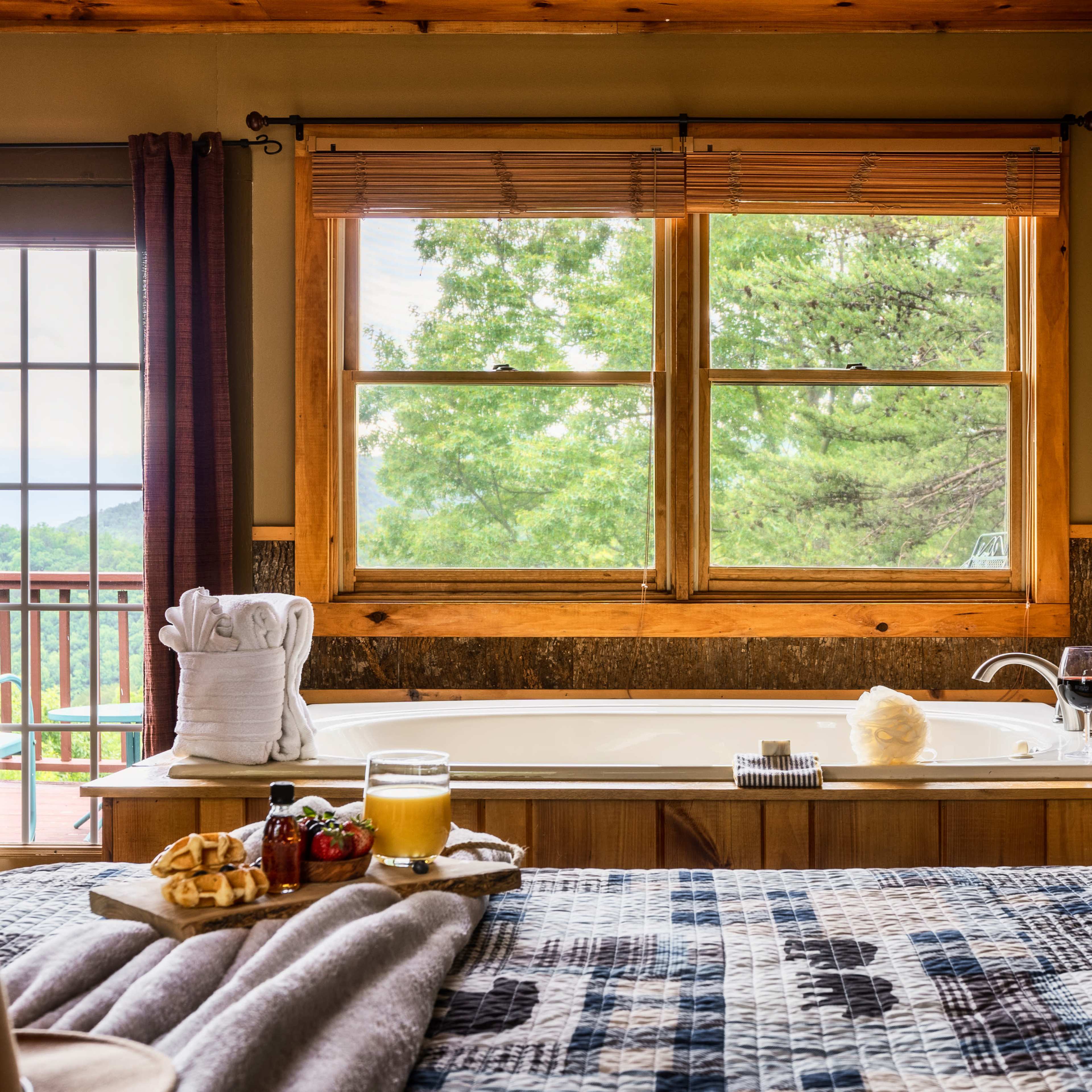 A cozy bedroom features a large window overlooking greenery, with a bathtub nearby and a tray of snacks on the bed.