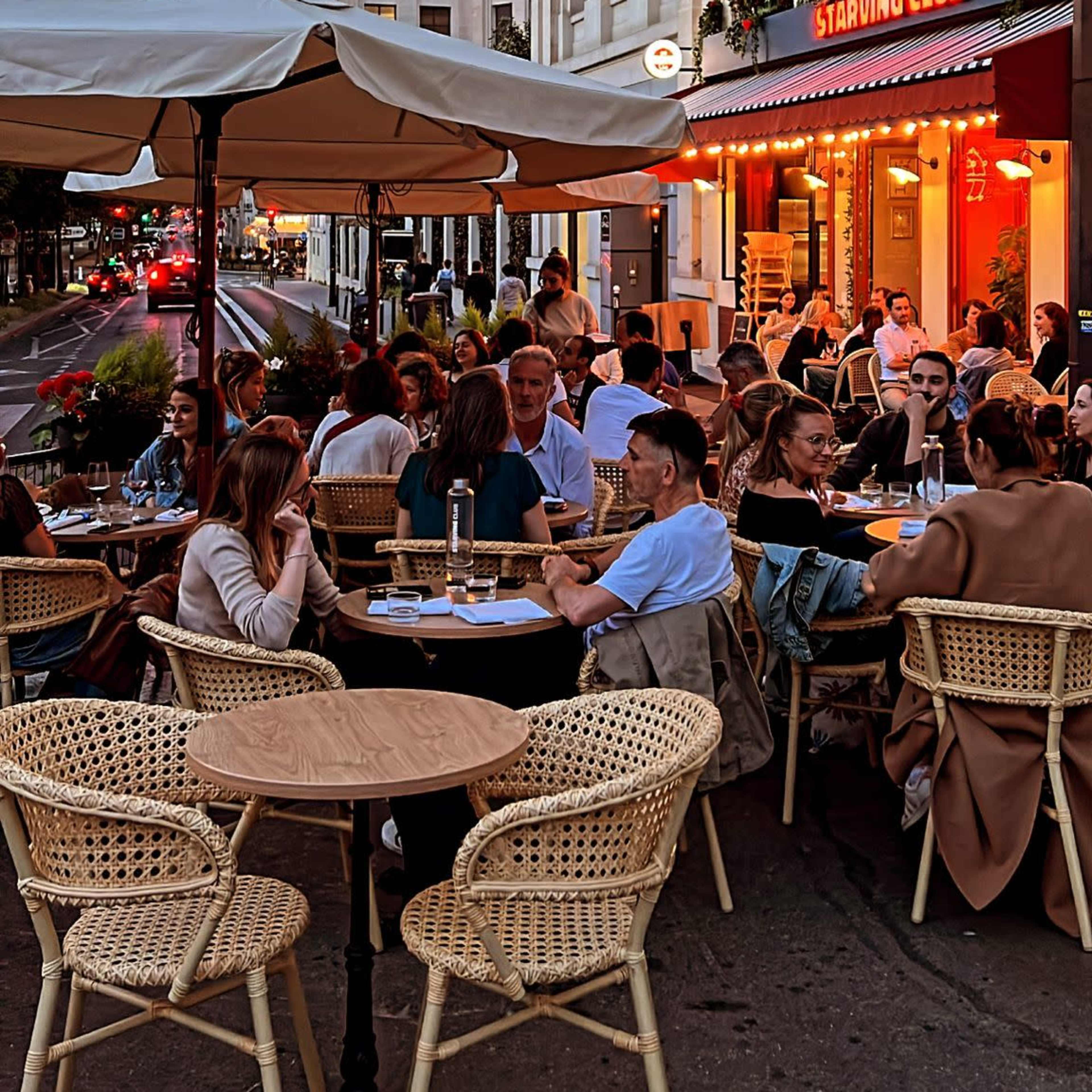 A busy street-side restaurant patio is filled with diners enjoying the evening atmosphere under string lights and nearby buildings.
