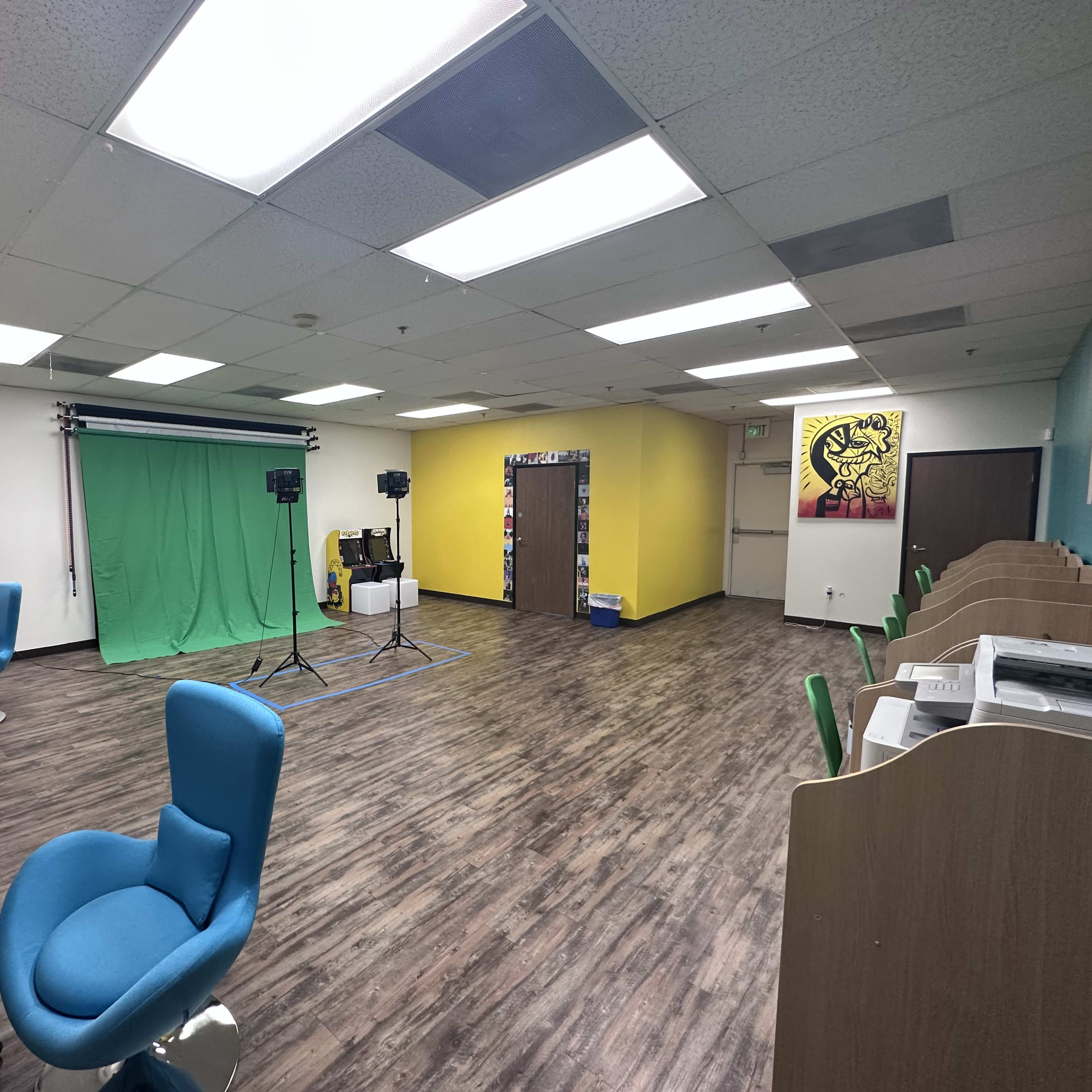 A spacious room with a green screen setup, studio lights, and several blue and white chairs, along with a colorful yellow wall and shelves in the background.