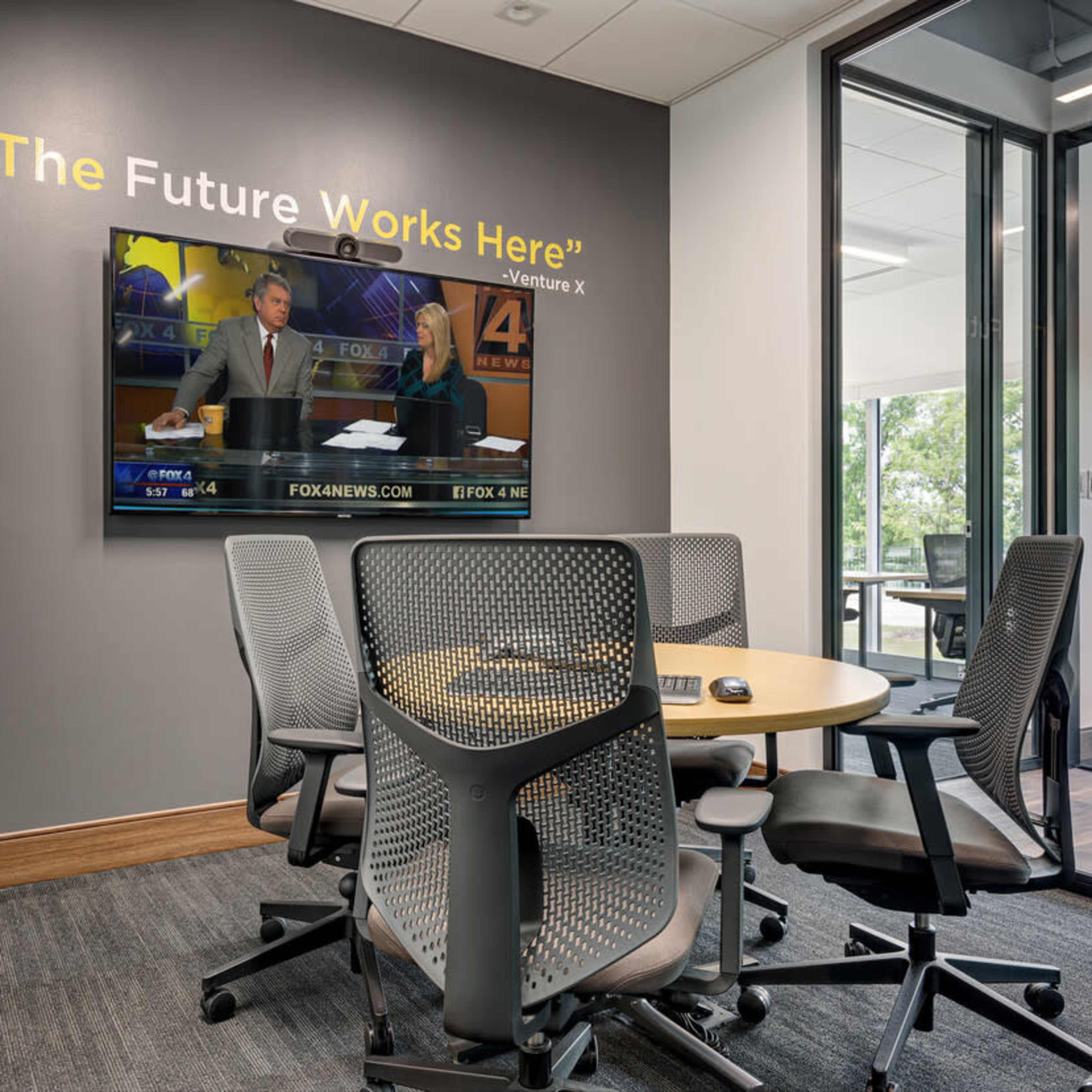 A conference room features a round table surrounded by ergonomic chairs, with a television displaying a news broadcast mounted on the wall.
