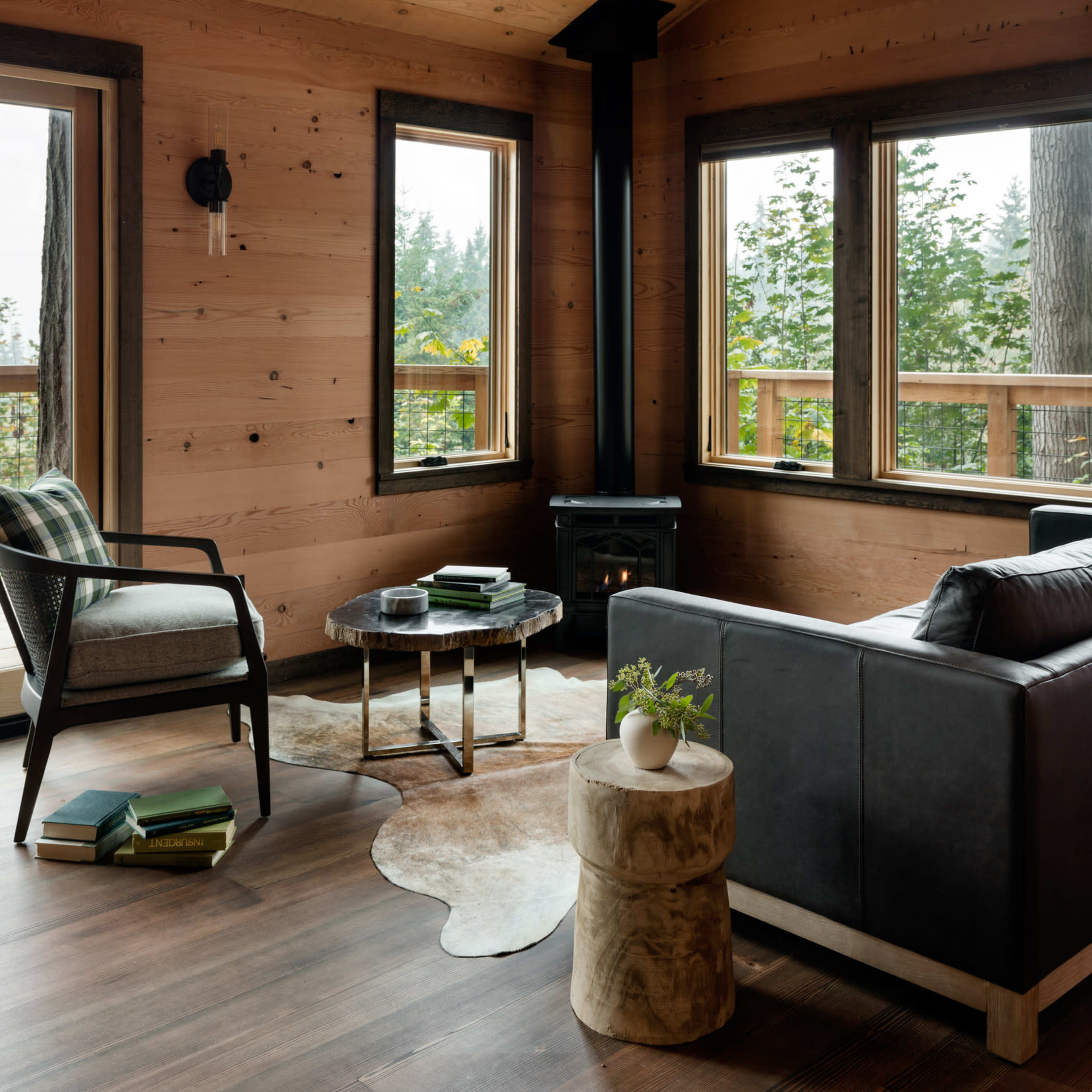 A cozy living room with wooden walls, a modern black sofa, a round coffee table, and large windows overlooking greenery.