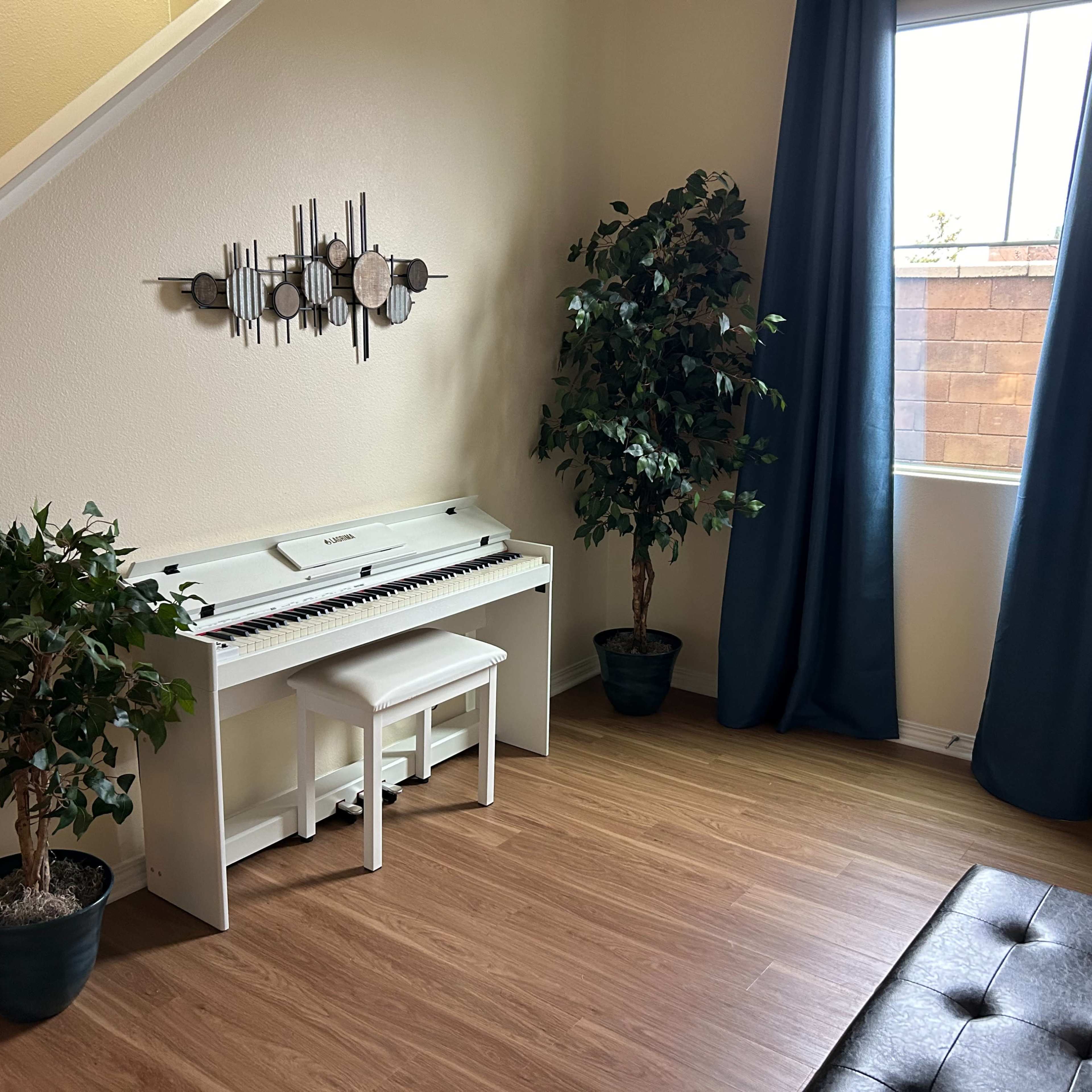 A white piano sits beside two potted plants in a room with wood flooring and a window covered by dark curtains.