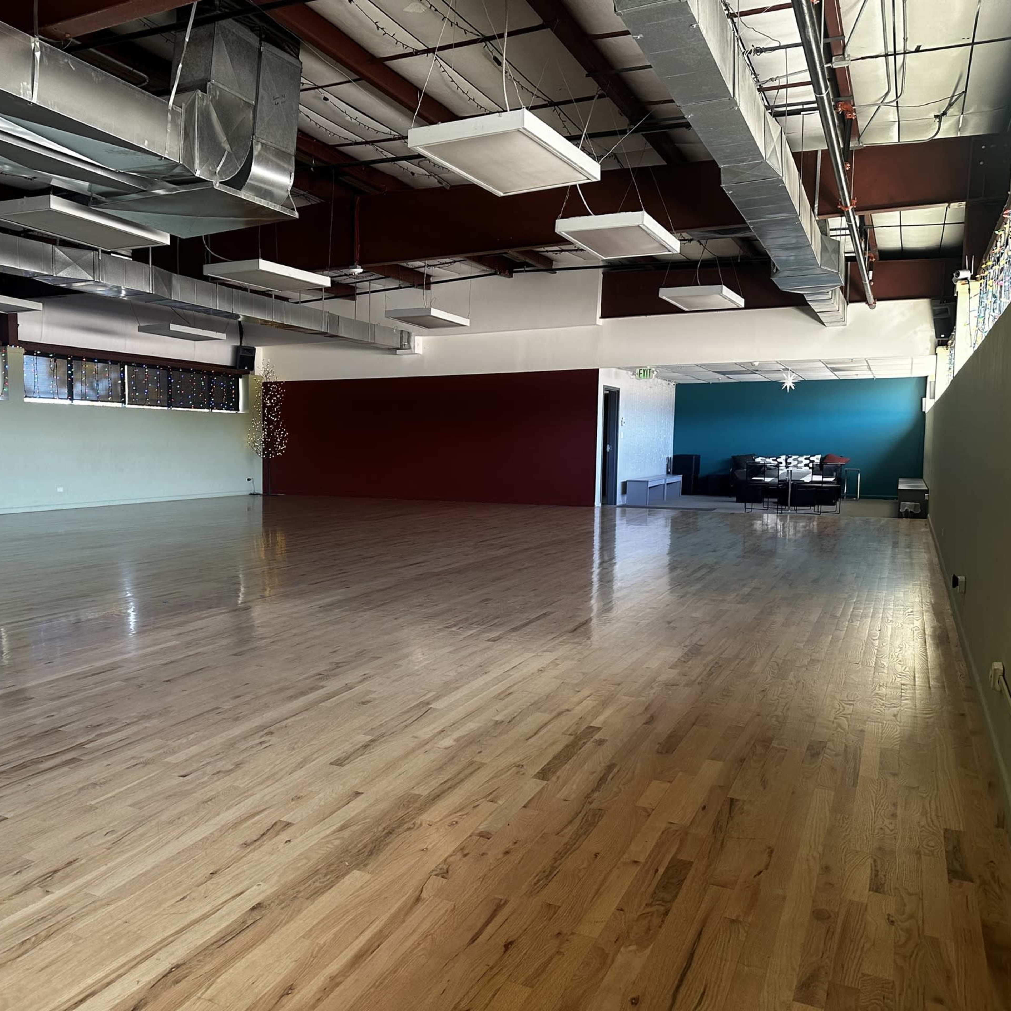 A spacious, empty studio with polished wooden floors and large windows, featuring a seating area in the back.