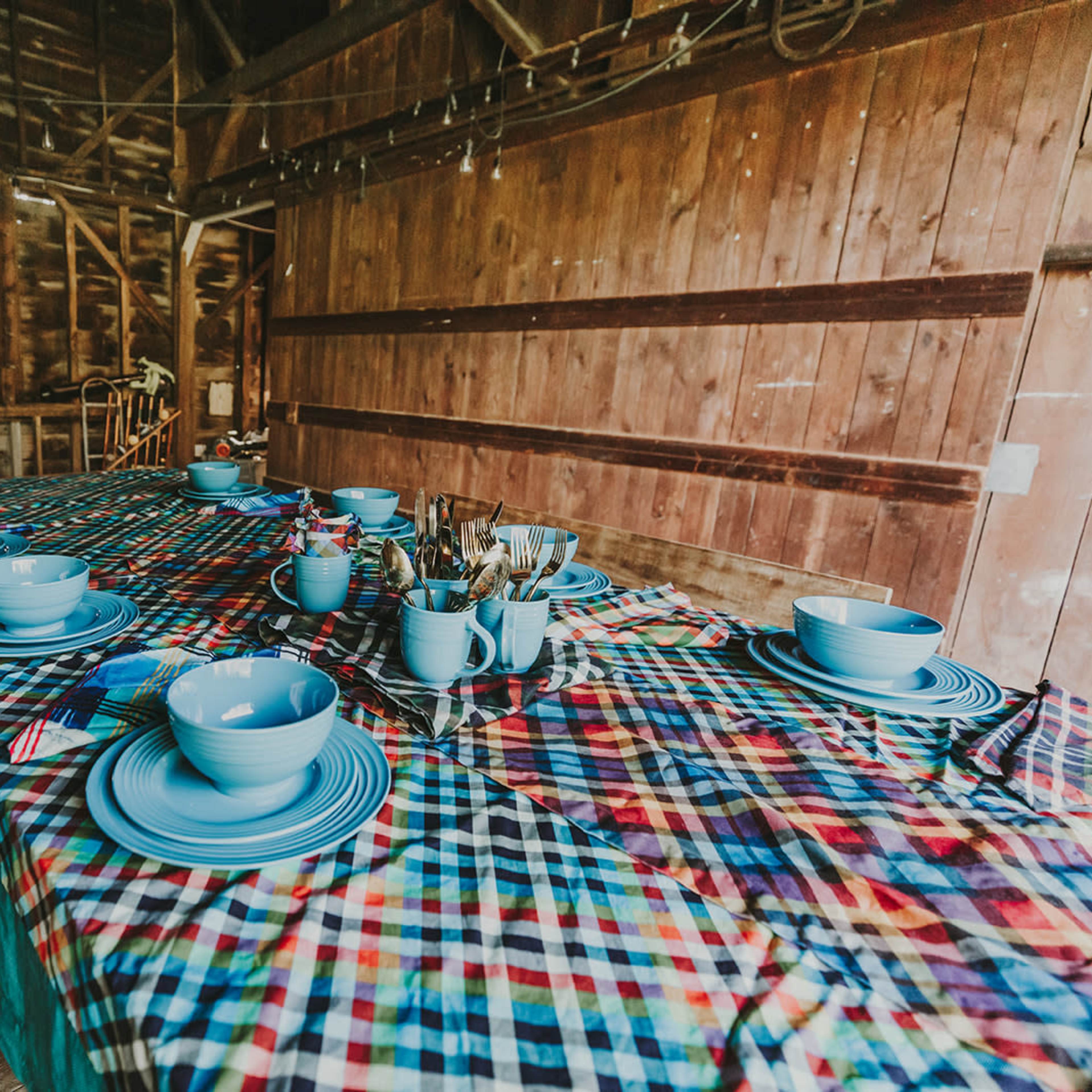 A long table covered with a plaid tablecloth is set with blue dishes and utensils in a rustic wooden interior.