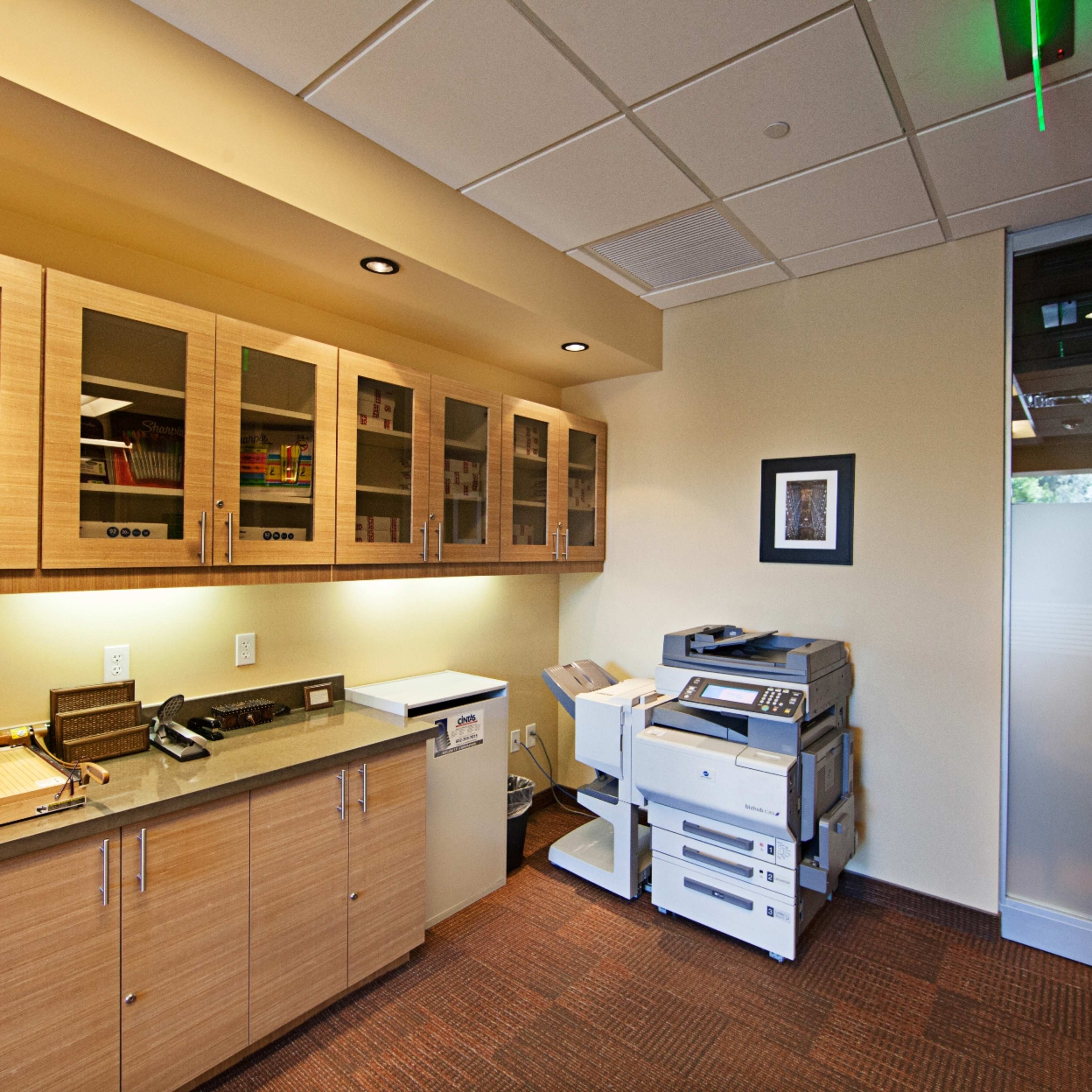 An office space with wooden cabinets, a countertop, and a multi-function printer near a glass partition.