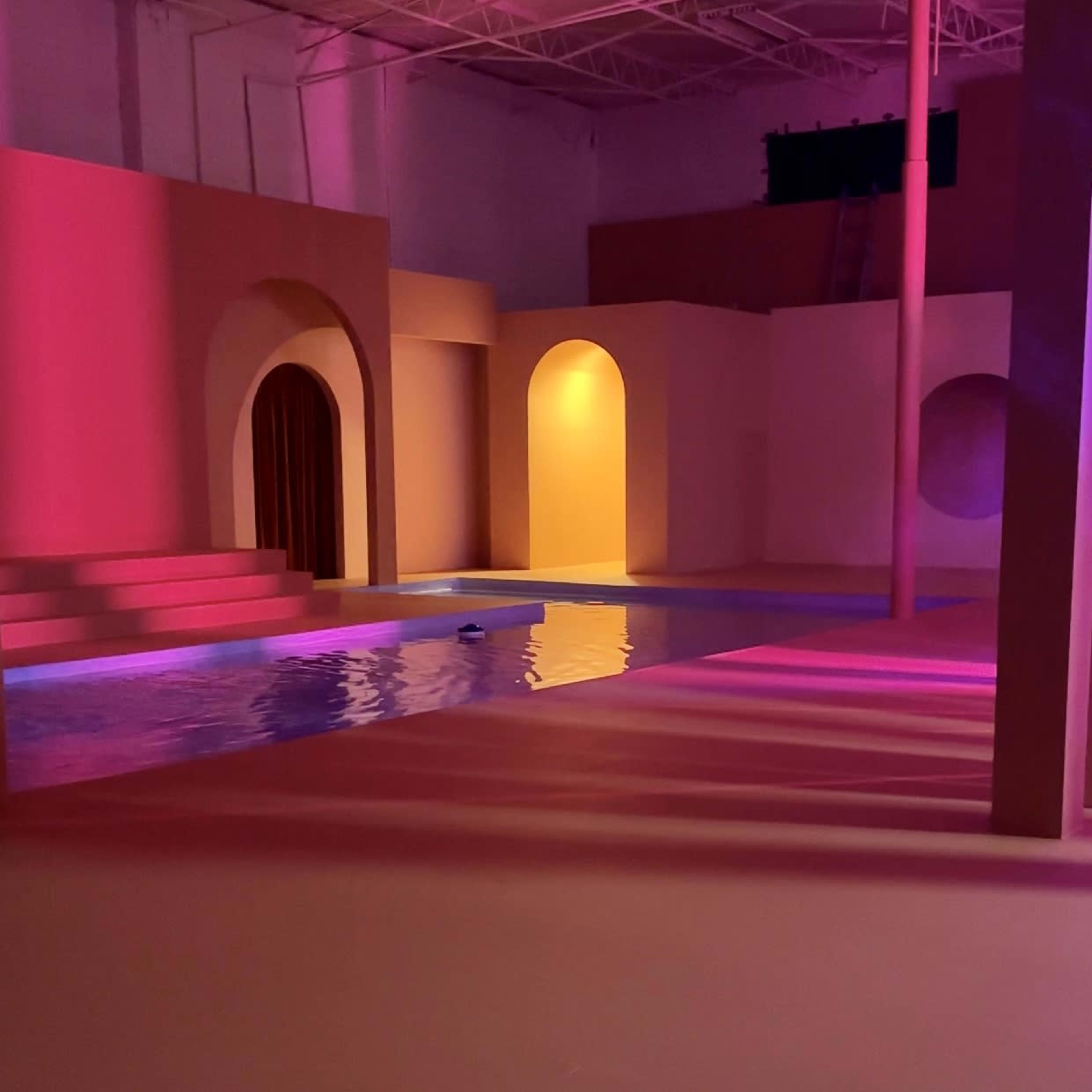 An indoor space featuring geometric arches, a small pool of water, and colored lighting.