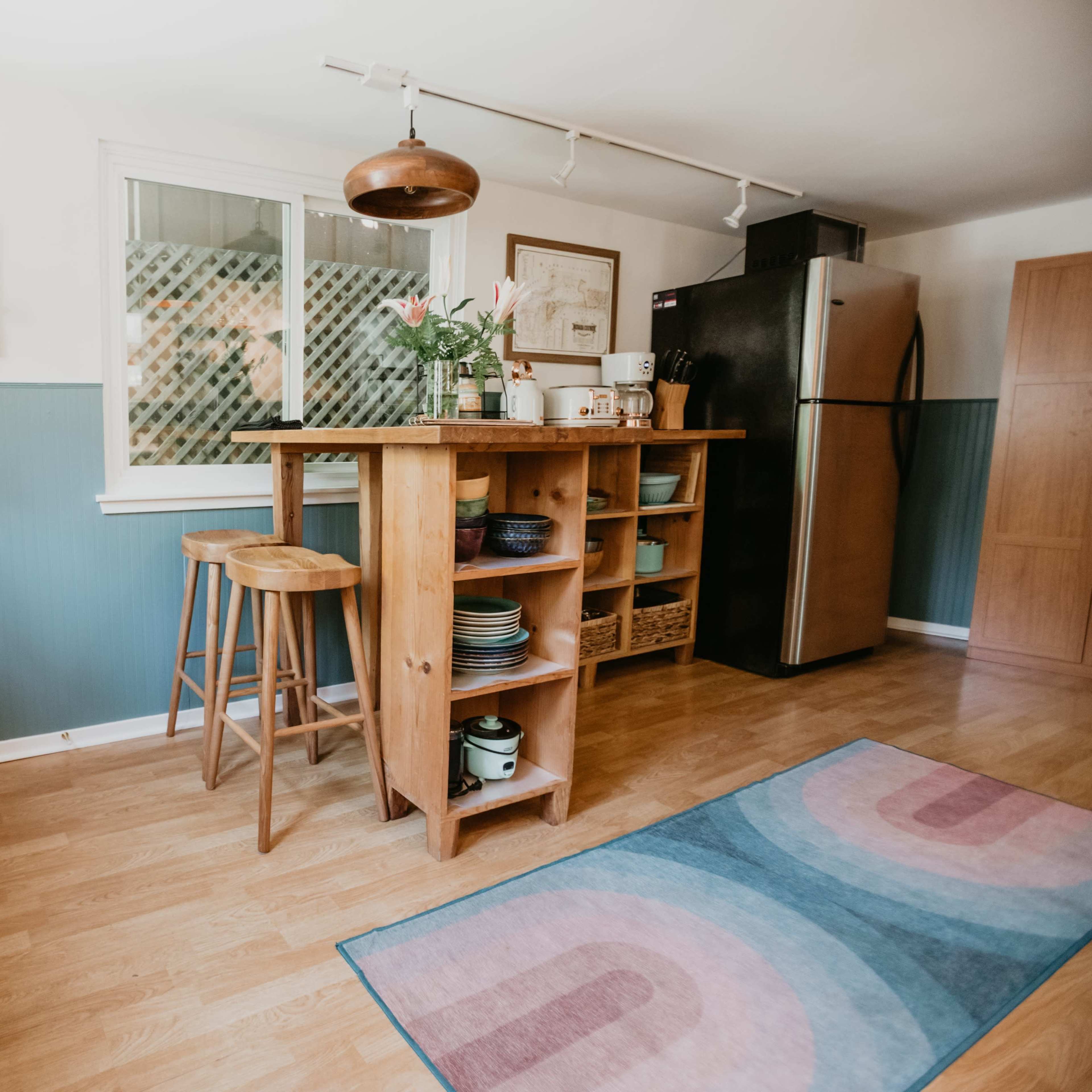 A cozy kitchen with a wooden bar, stools, and a refrigerator, featuring a door with a window and a colorful rug on the floor.