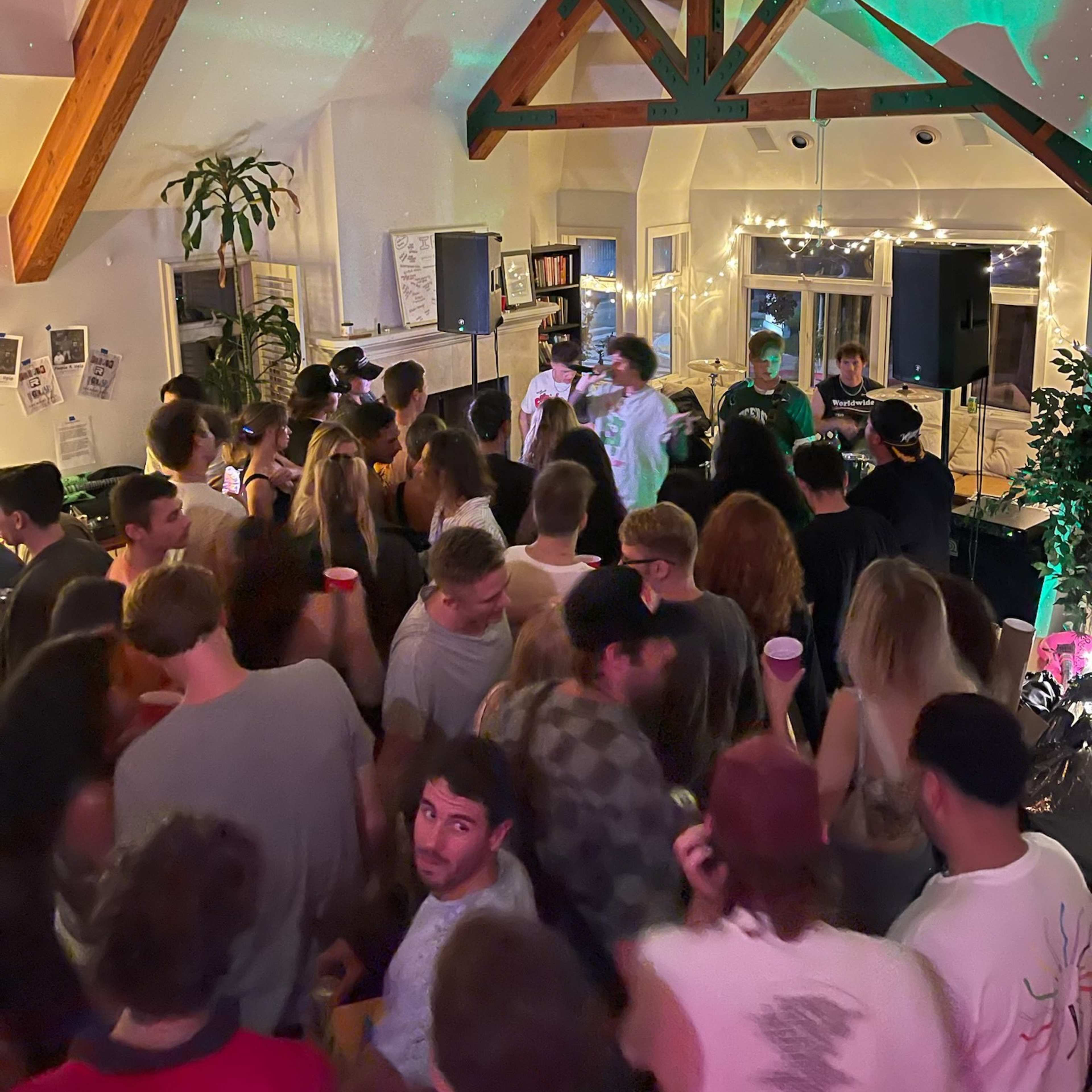 A crowd gathers in a decorated room with wooden beams, enjoying a live performance on a small stage.