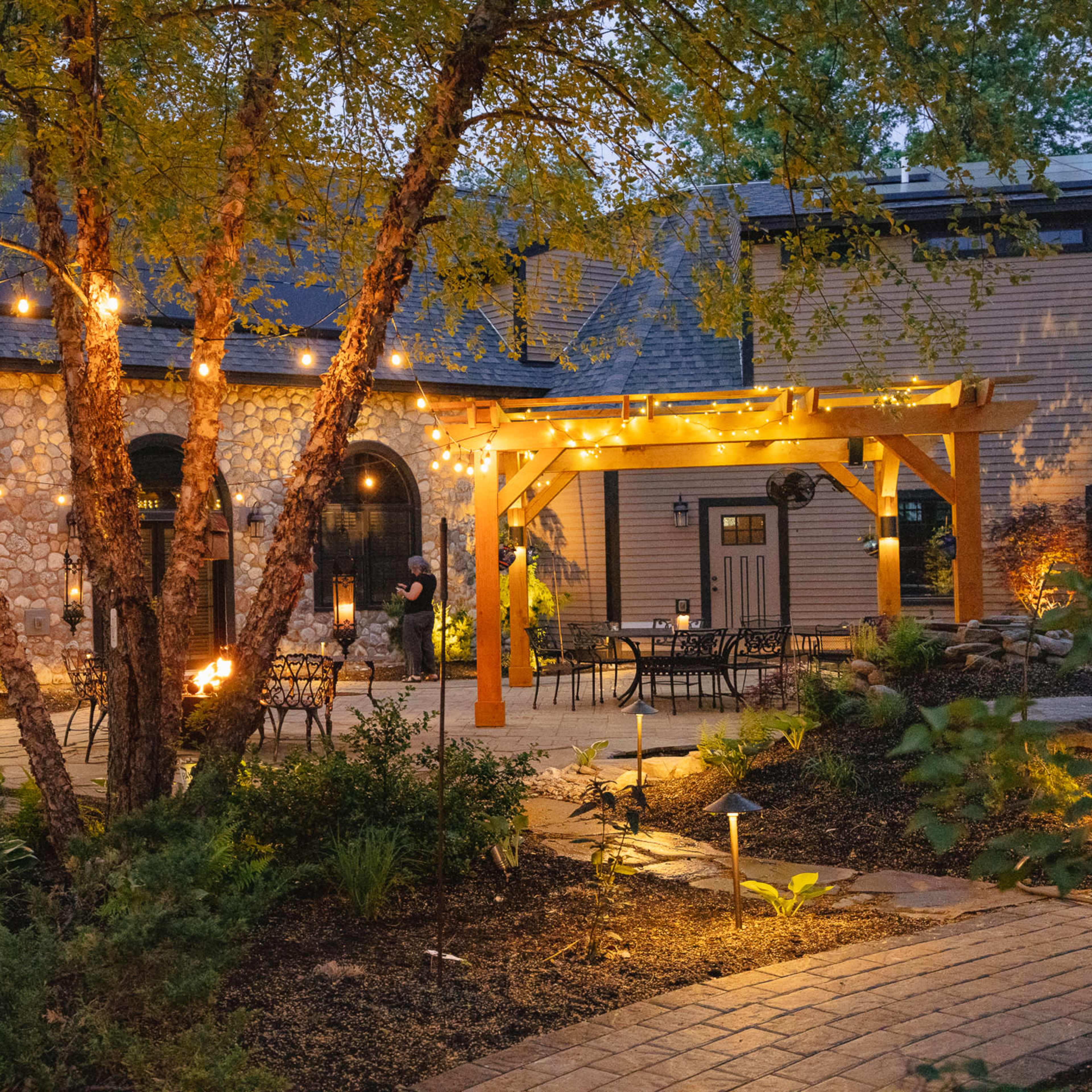 A stone building with arched windows is surrounded by trees and outdoor seating under string lights in a landscaped courtyard.