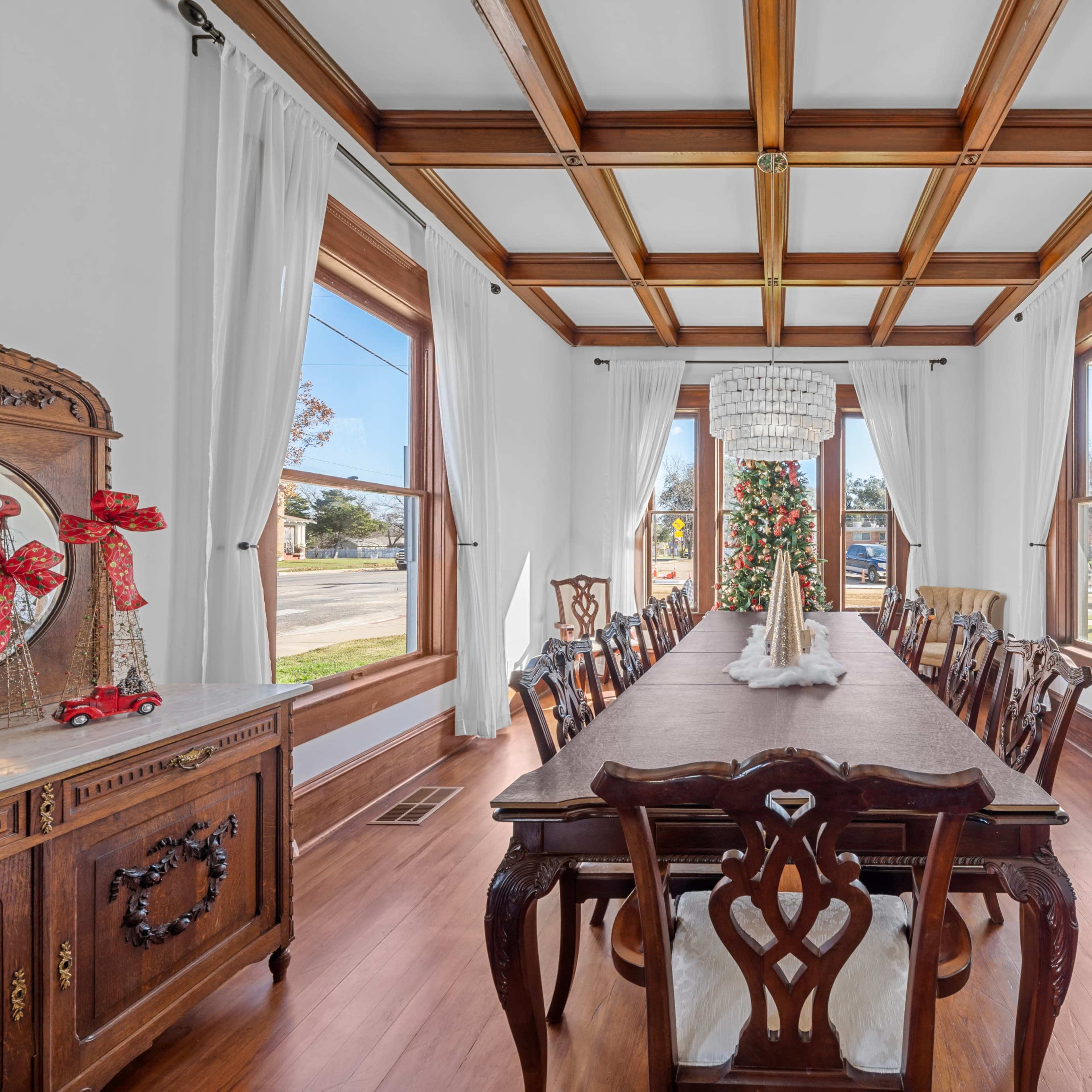 A long dining table with wooden chairs is set in a room featuring large windows and a coffered ceiling.