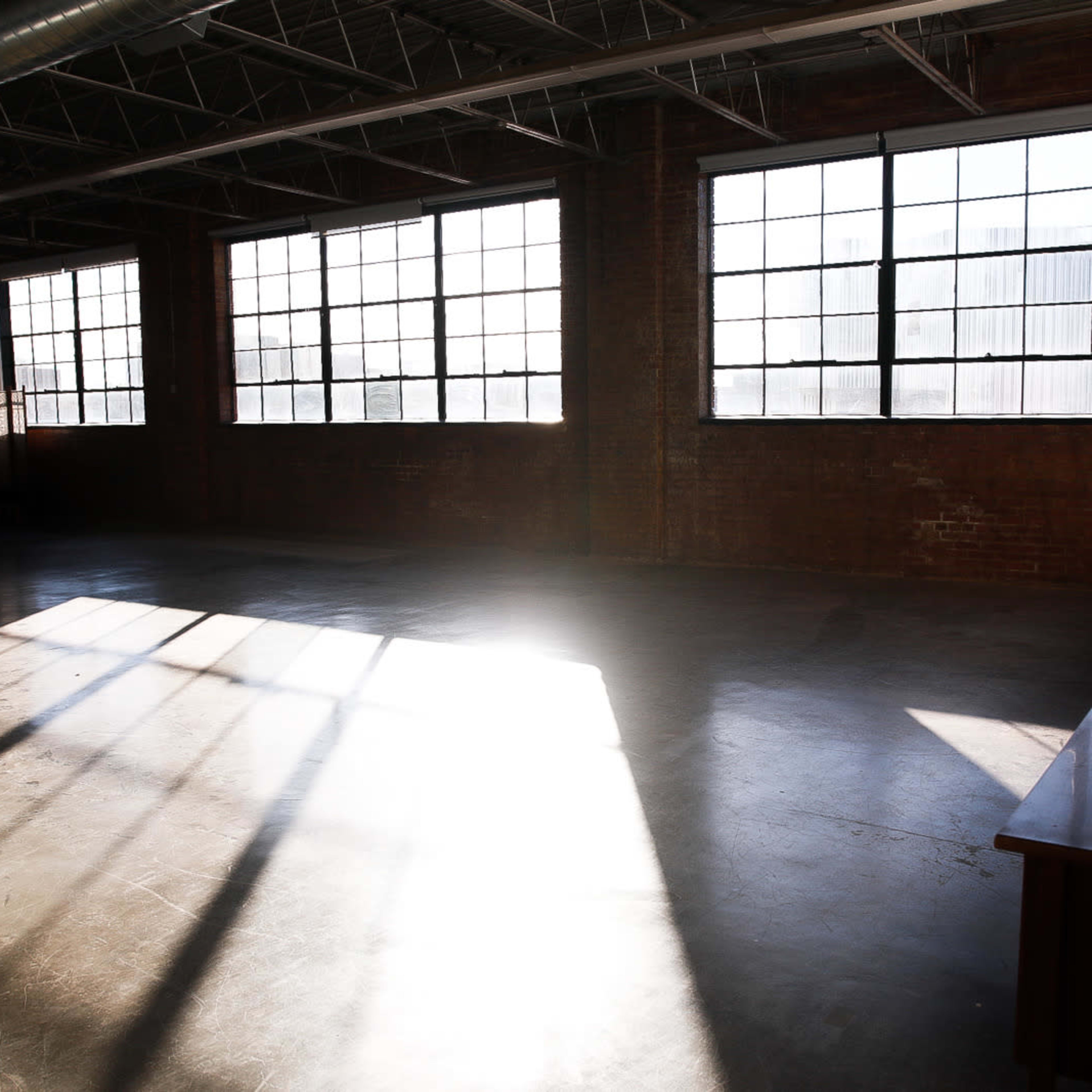 A large, empty room with concrete flooring and sunlight streaming through large industrial windows, casting shadows on the walls.