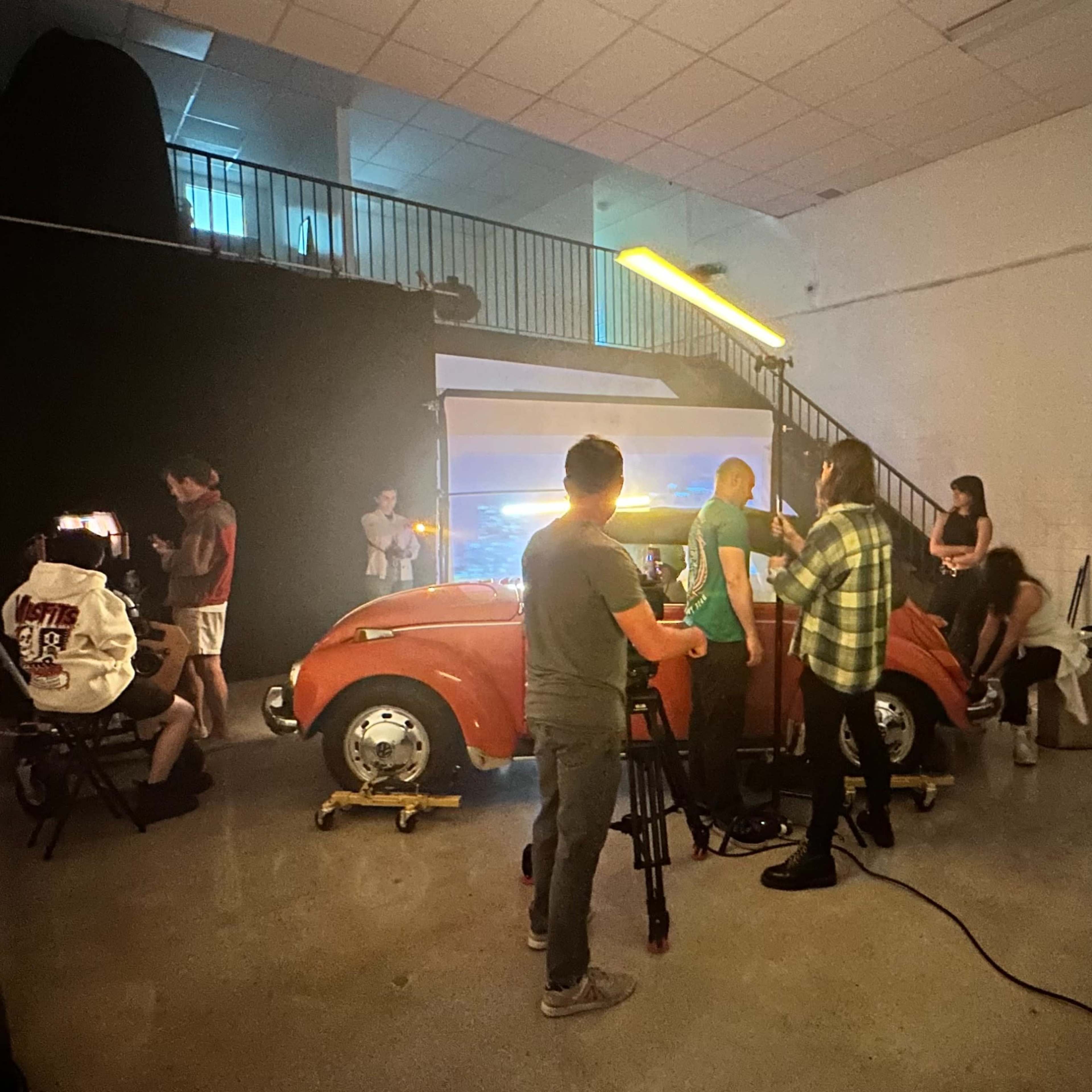 A group of people is filming a red vintage car inside a studio equipped with lighting and cameras, with additional crew members setting up in the background.