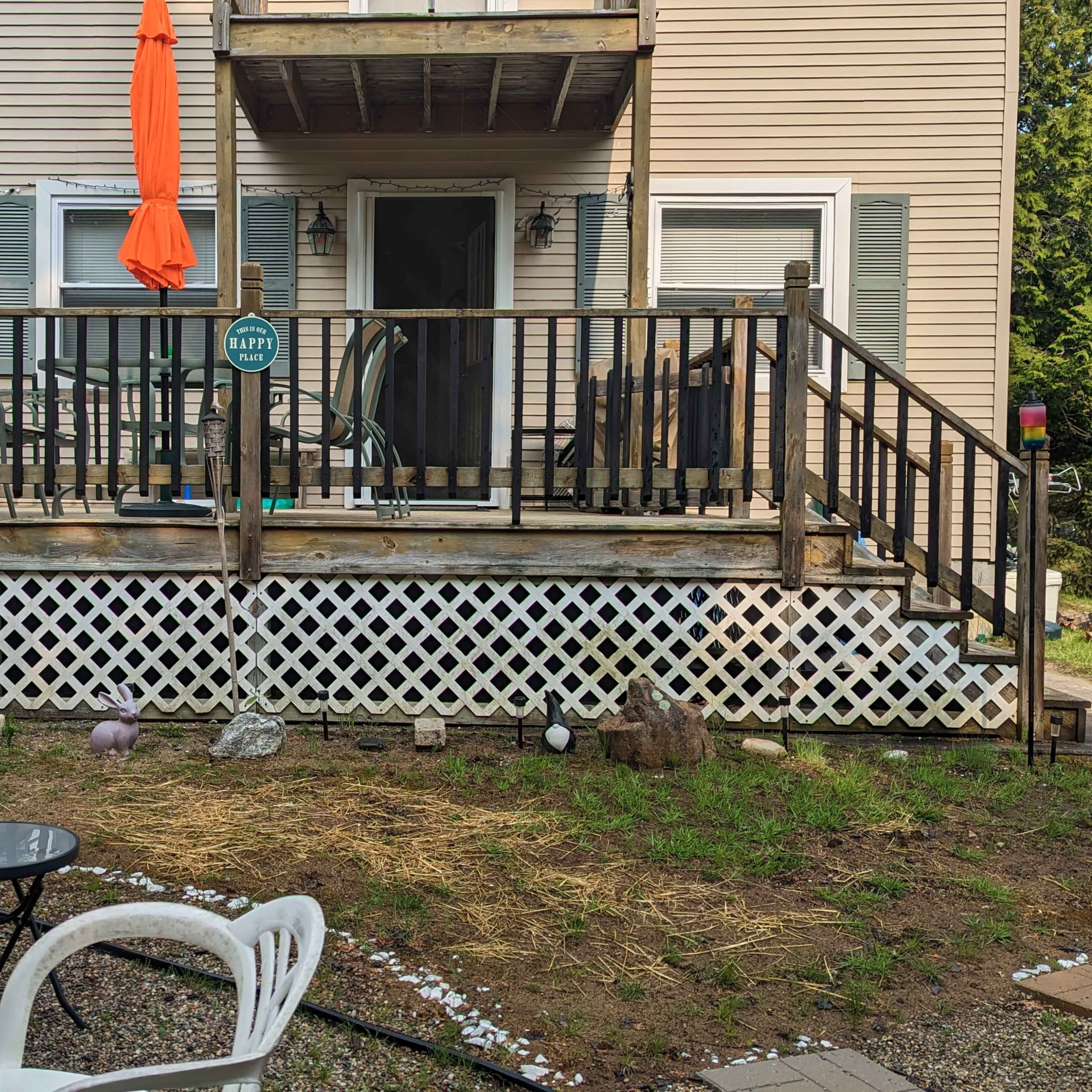 A wooden deck with chairs and an orange umbrella in front of a two-story house, surrounded by a lawn with patchy grass and gravel.