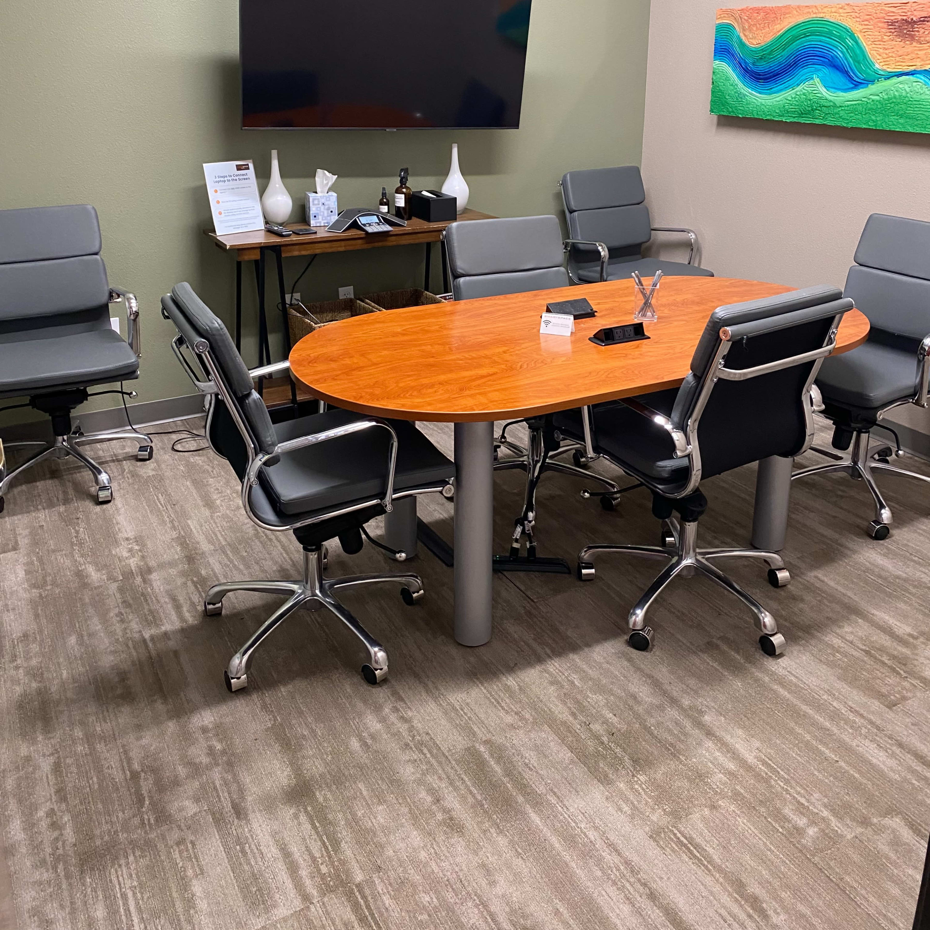 A conference room with a wooden oval table surrounded by six gray chairs, a large wall-mounted TV, and an abstract painting on the wall.