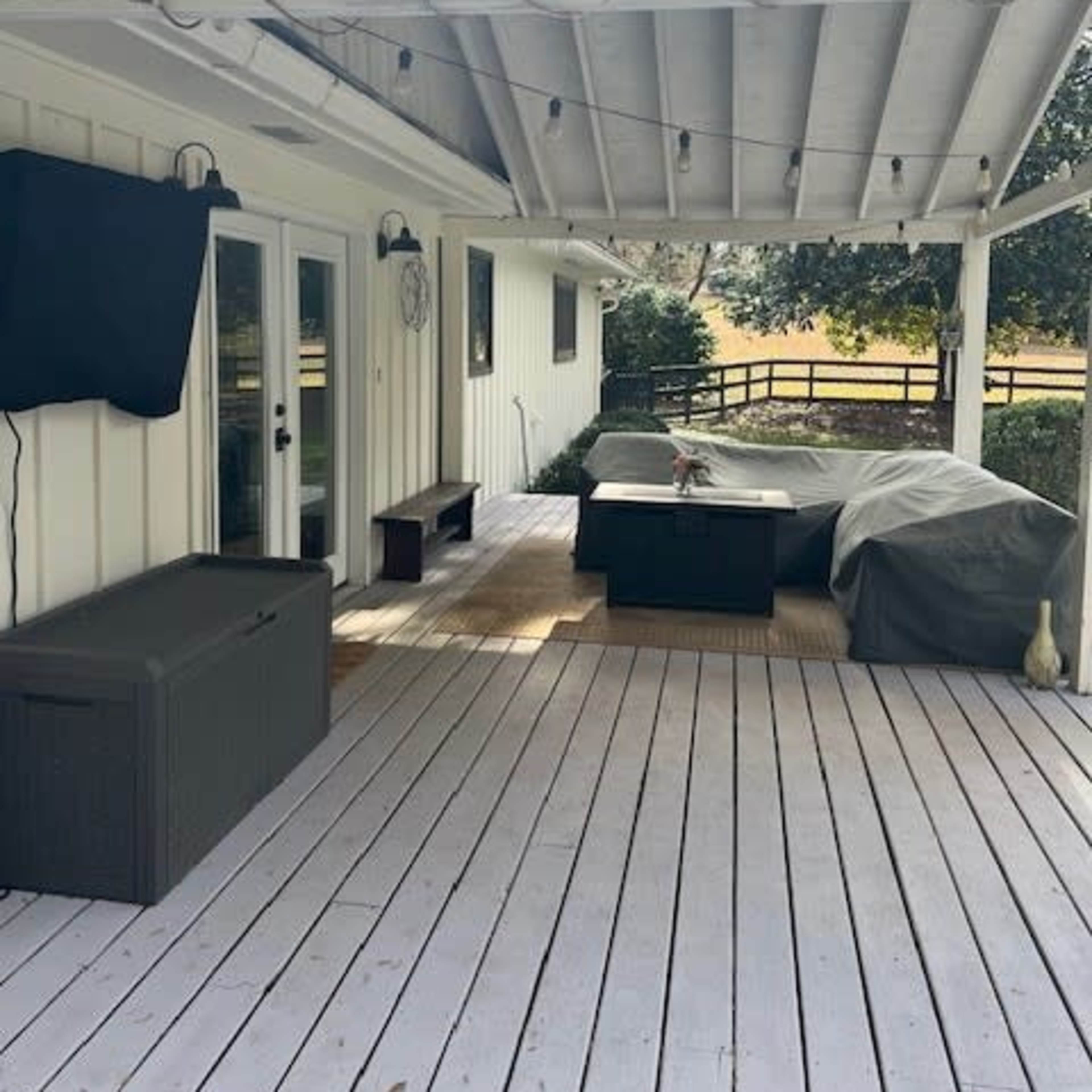 A covered outdoor patio features a dining table with a protective cover, a storage bench, and a mounted television against a white wall.