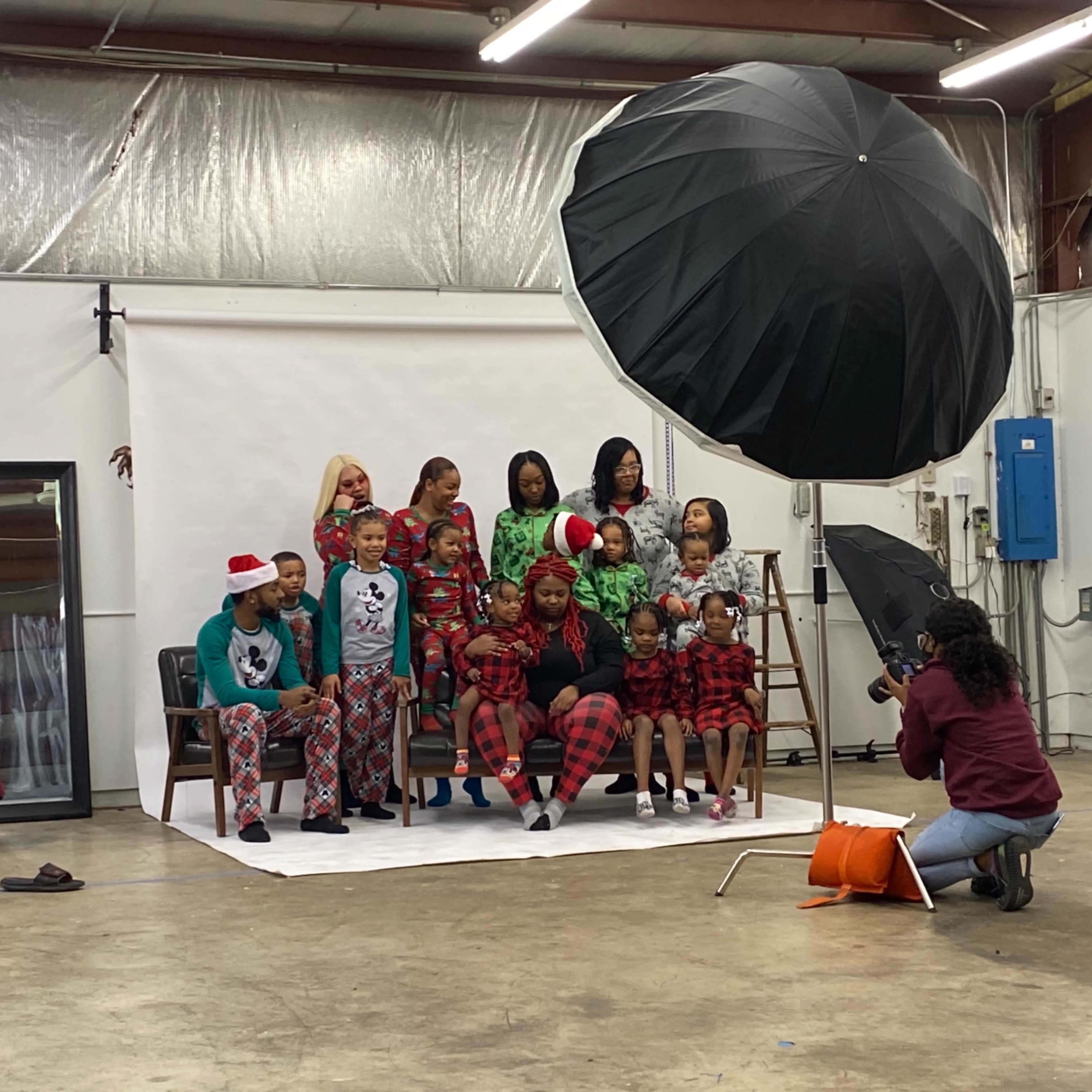 A group of people wearing matching holiday pajamas poses for a family photo in a studio setting with a large umbrella light.