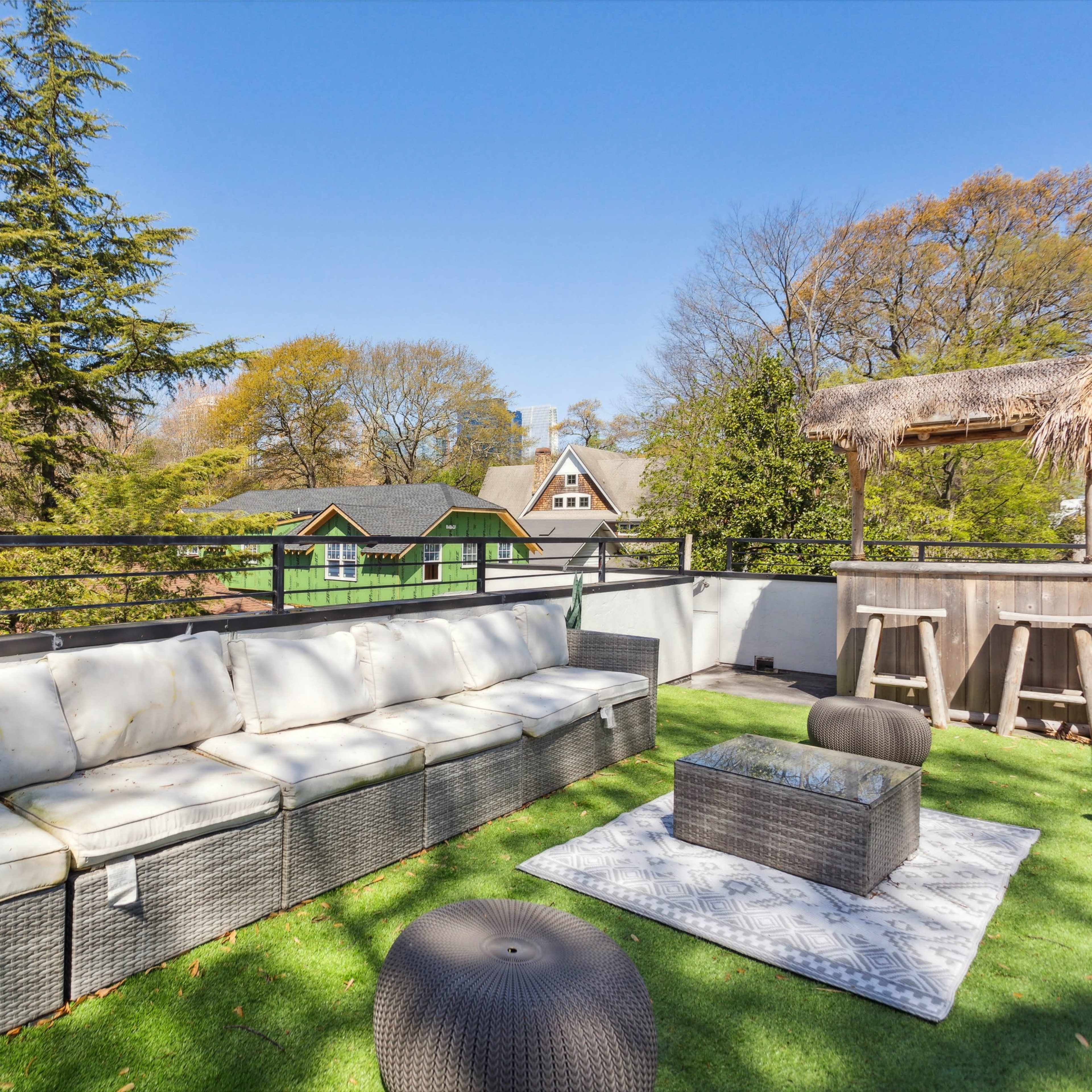 A rooftop terrace features a large sectional sofa, a decorative area rug, and a bar area surrounded by trees and rooftops.