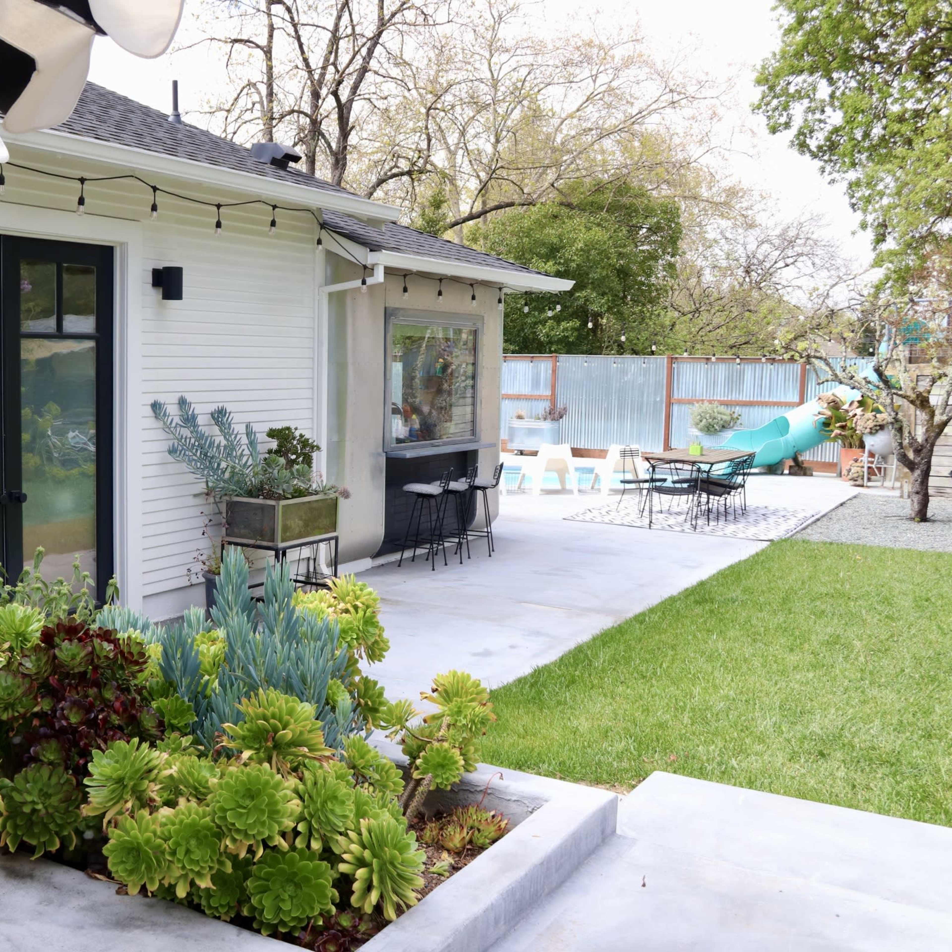 A modern backyard with a plant-filled garden, patio area, and outdoor seating.