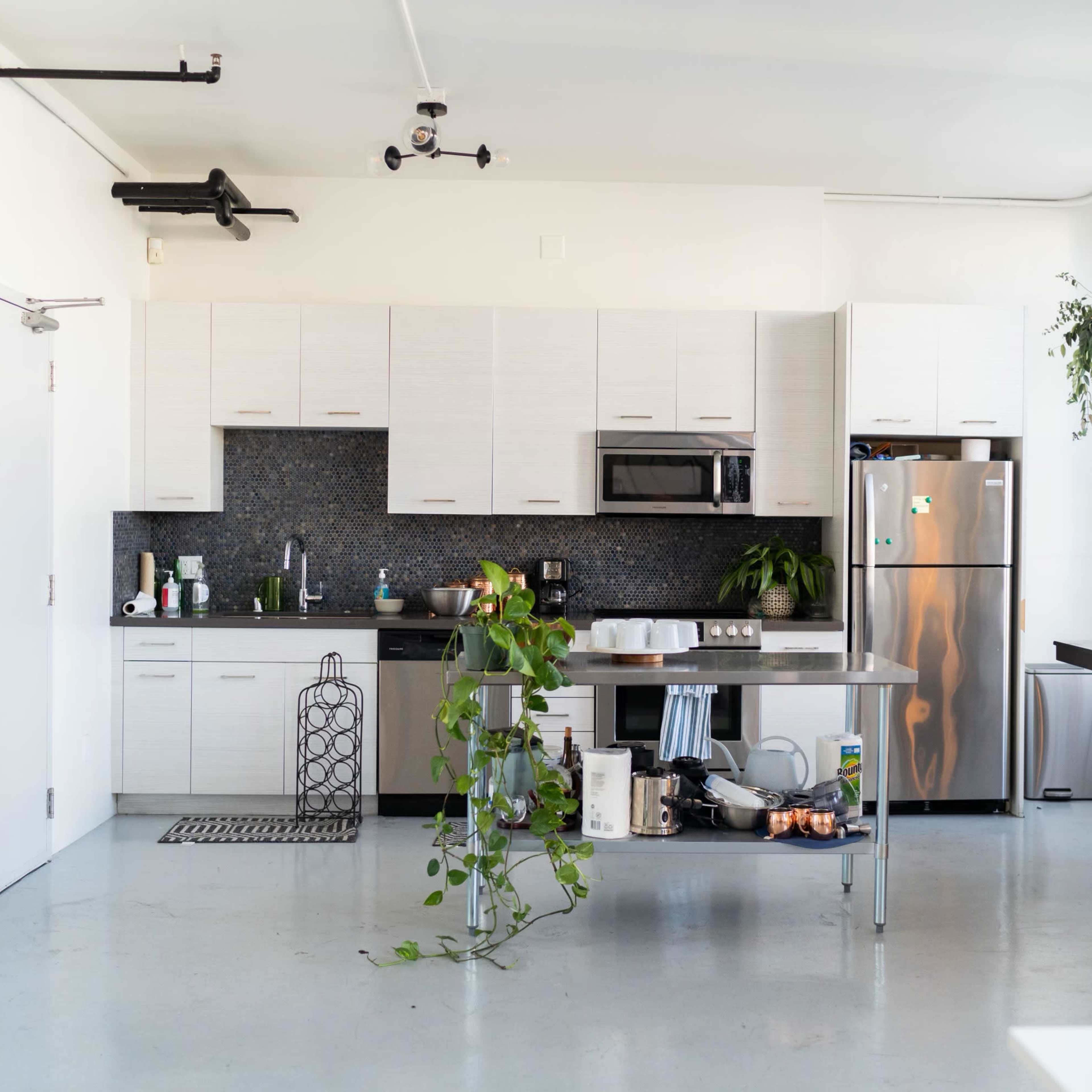 A modern kitchen and dining area features sleek cabinetry, a stainless steel refrigerator, and a table with chairs, accompanied by plants and decorative elements.