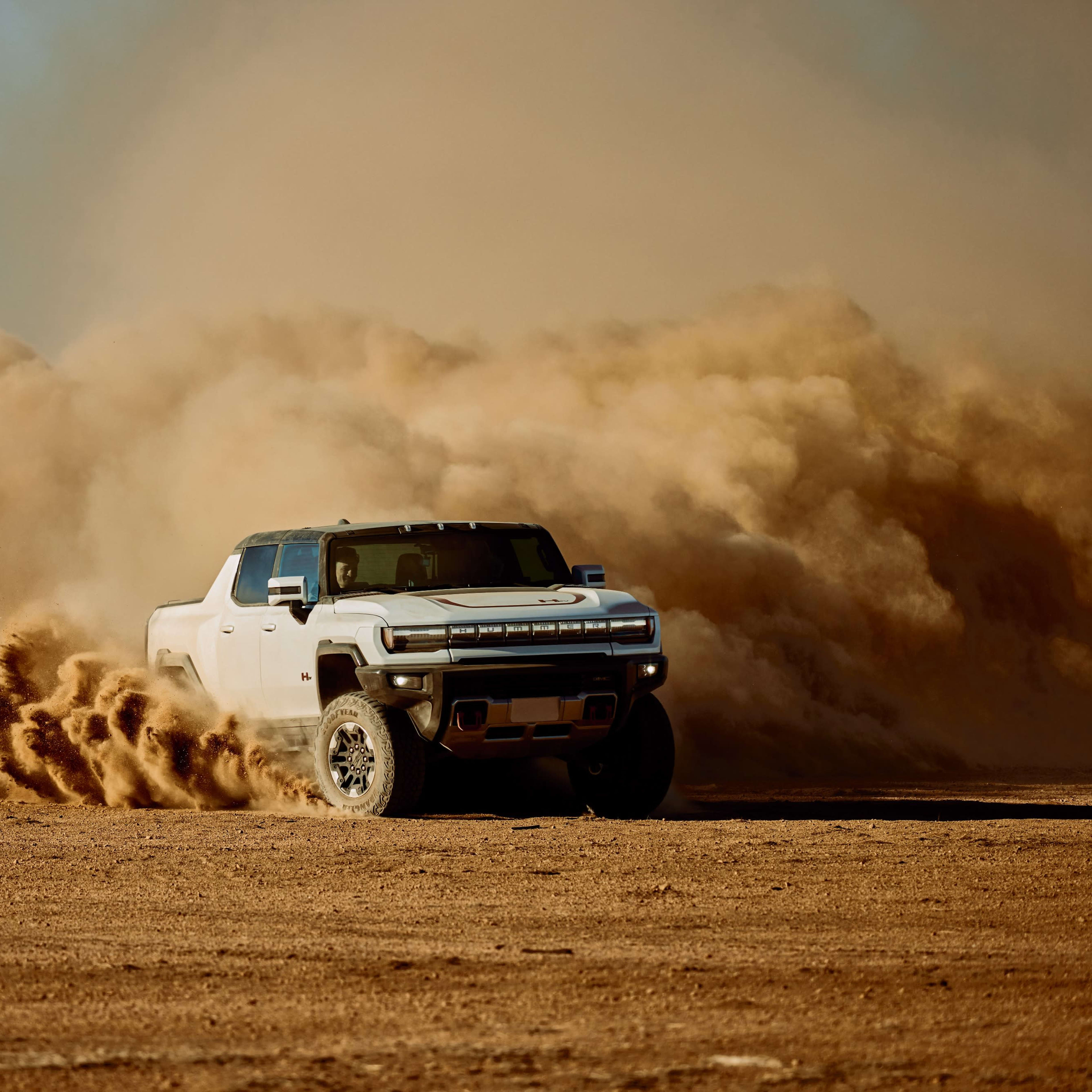 A white pickup truck kicks up a cloud of dust while driving on a dirt surface in a desert environment.