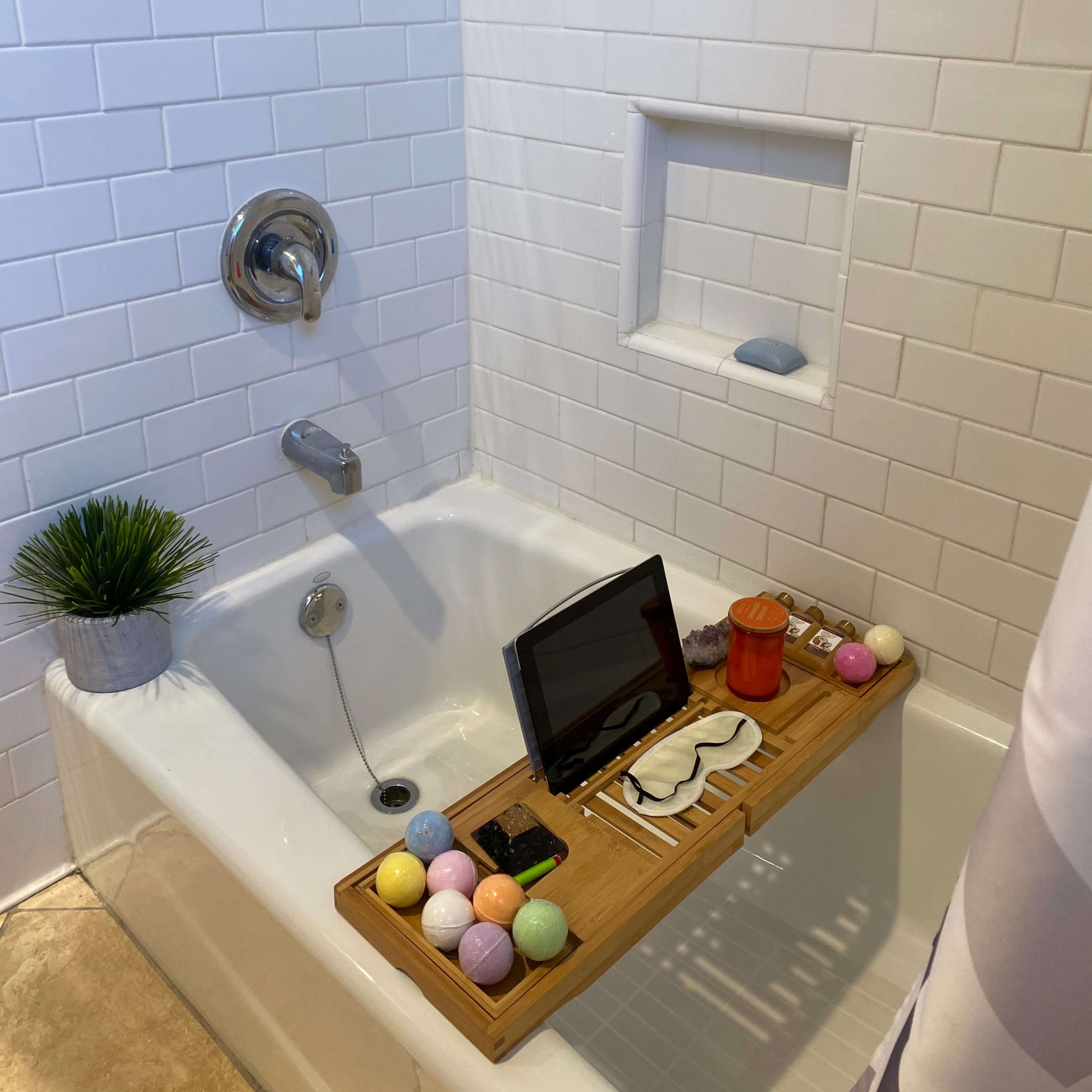 A clean, white tiled bathroom features a soaking tub adorned with a wooden tray holding various items, including an electronic device and colorful bath accessories.