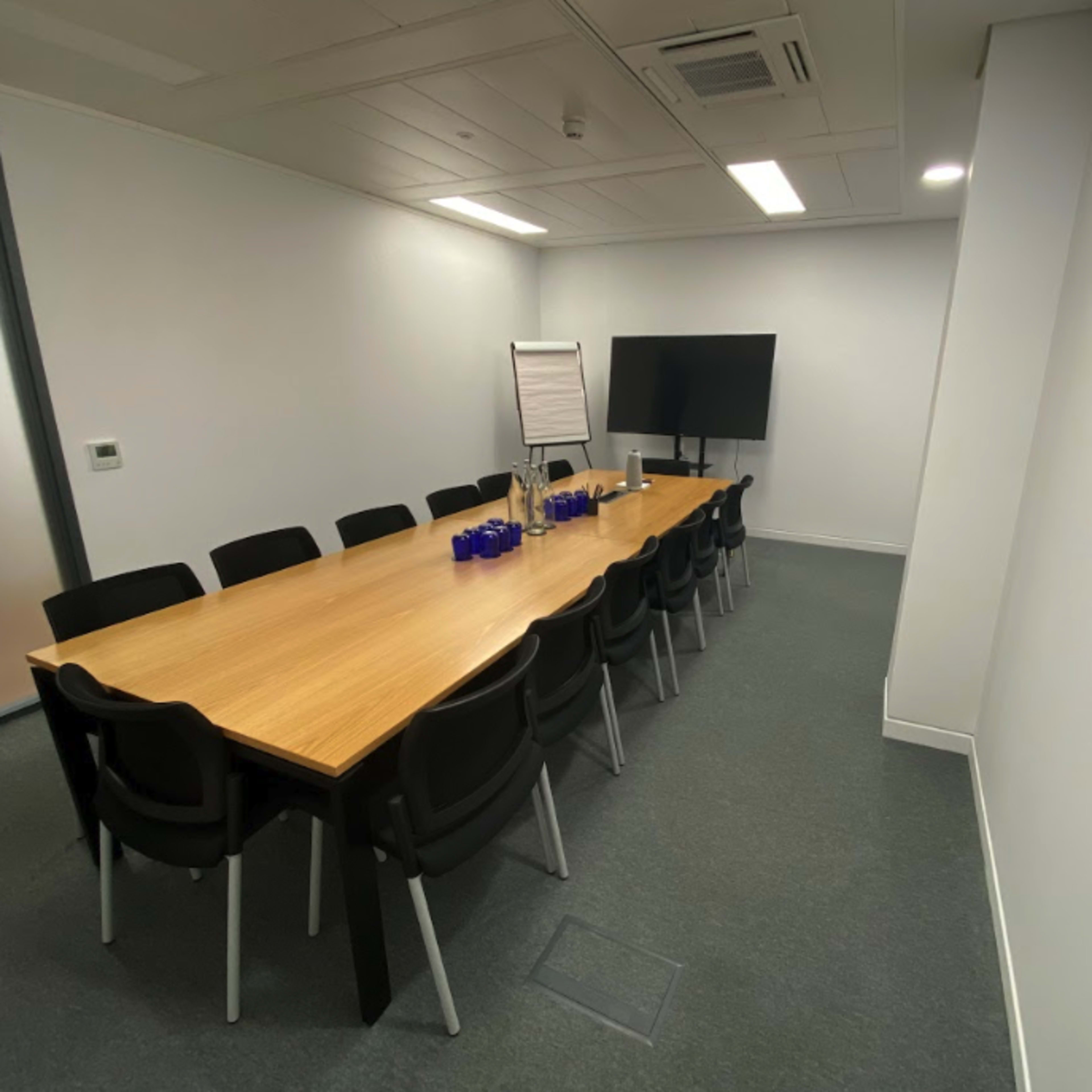 A conference room features a long wooden table surrounded by black chairs, with a whiteboard and a large screen on the walls.