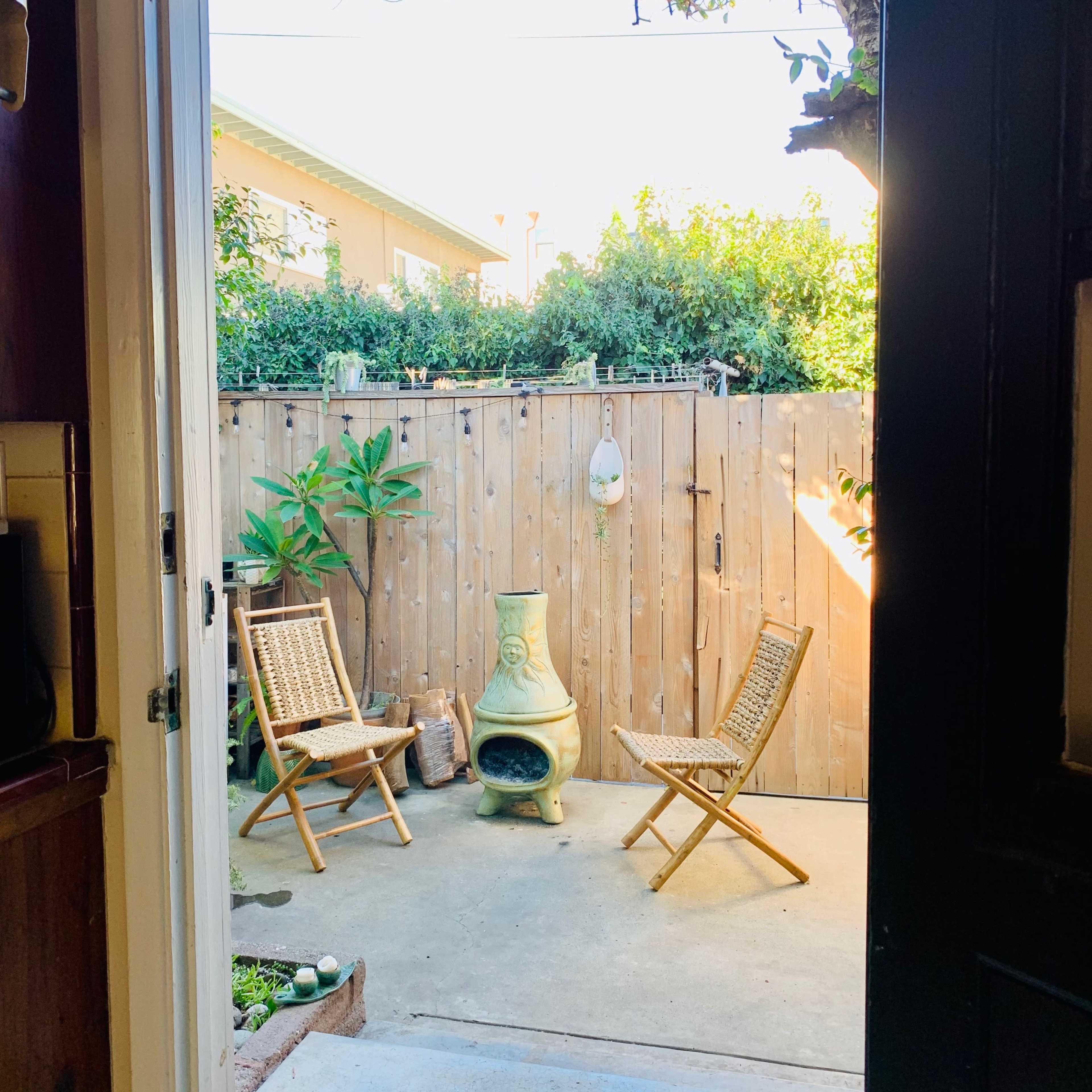 A view from an open door into a small outdoor patio with two wicker chairs and a green chiminea, surrounded by a wooden fence and plants.