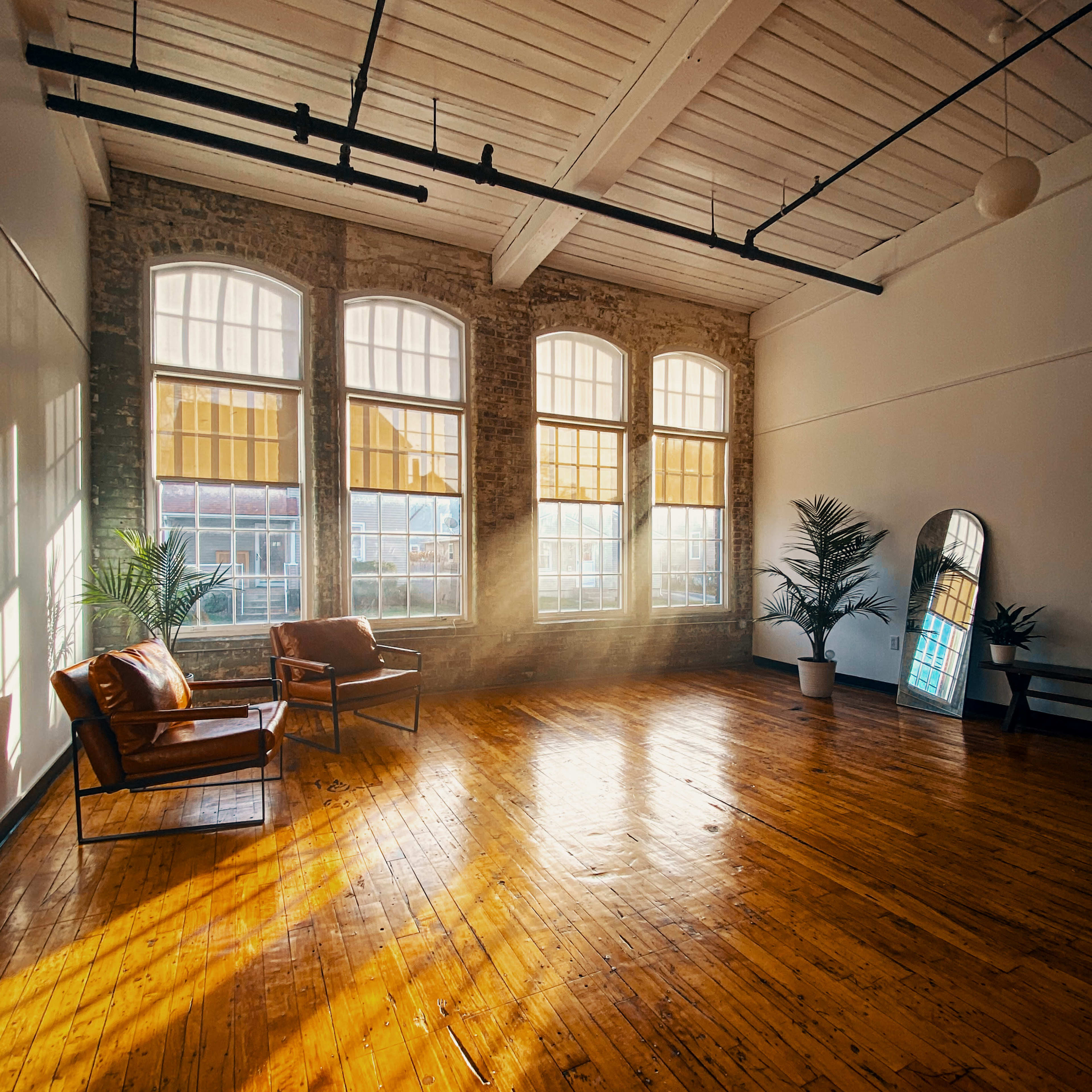 A spacious room with large windows, wooden flooring, and two leather chairs, illuminated by sunlight streaming through the windows.