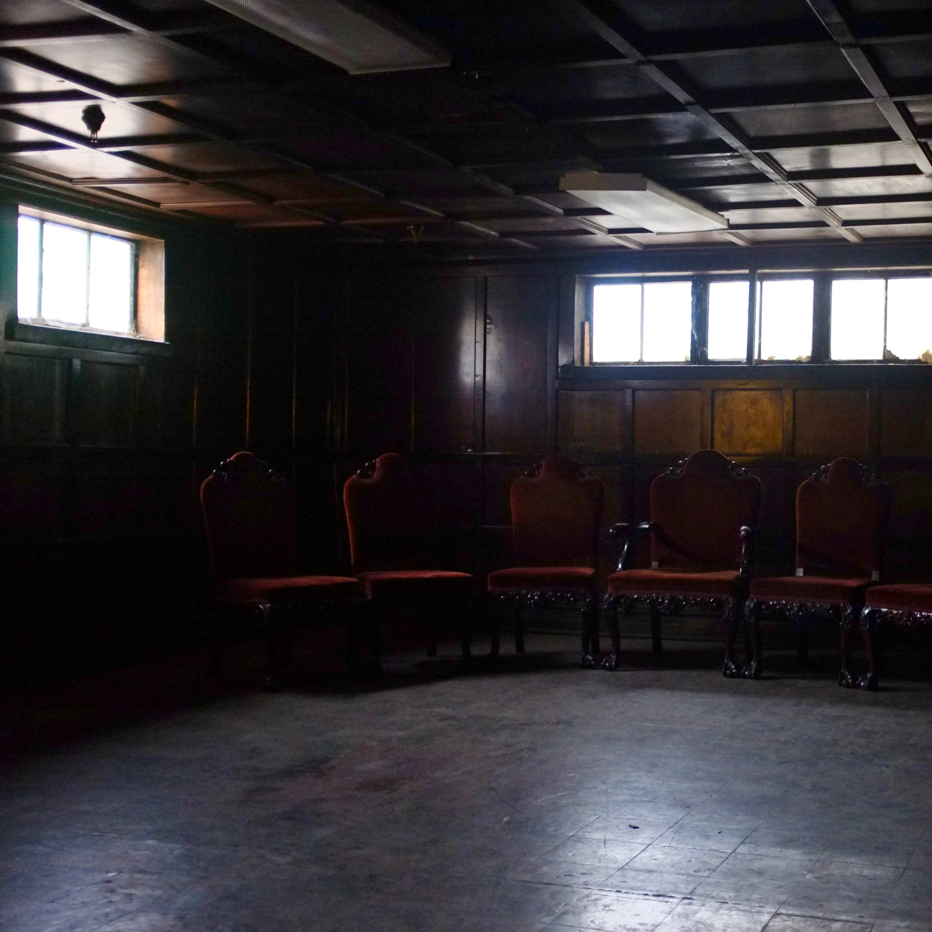 A dimly lit room features a row of five ornate chairs against a dark wooden wall, with two small windows allowing faint light to enter.