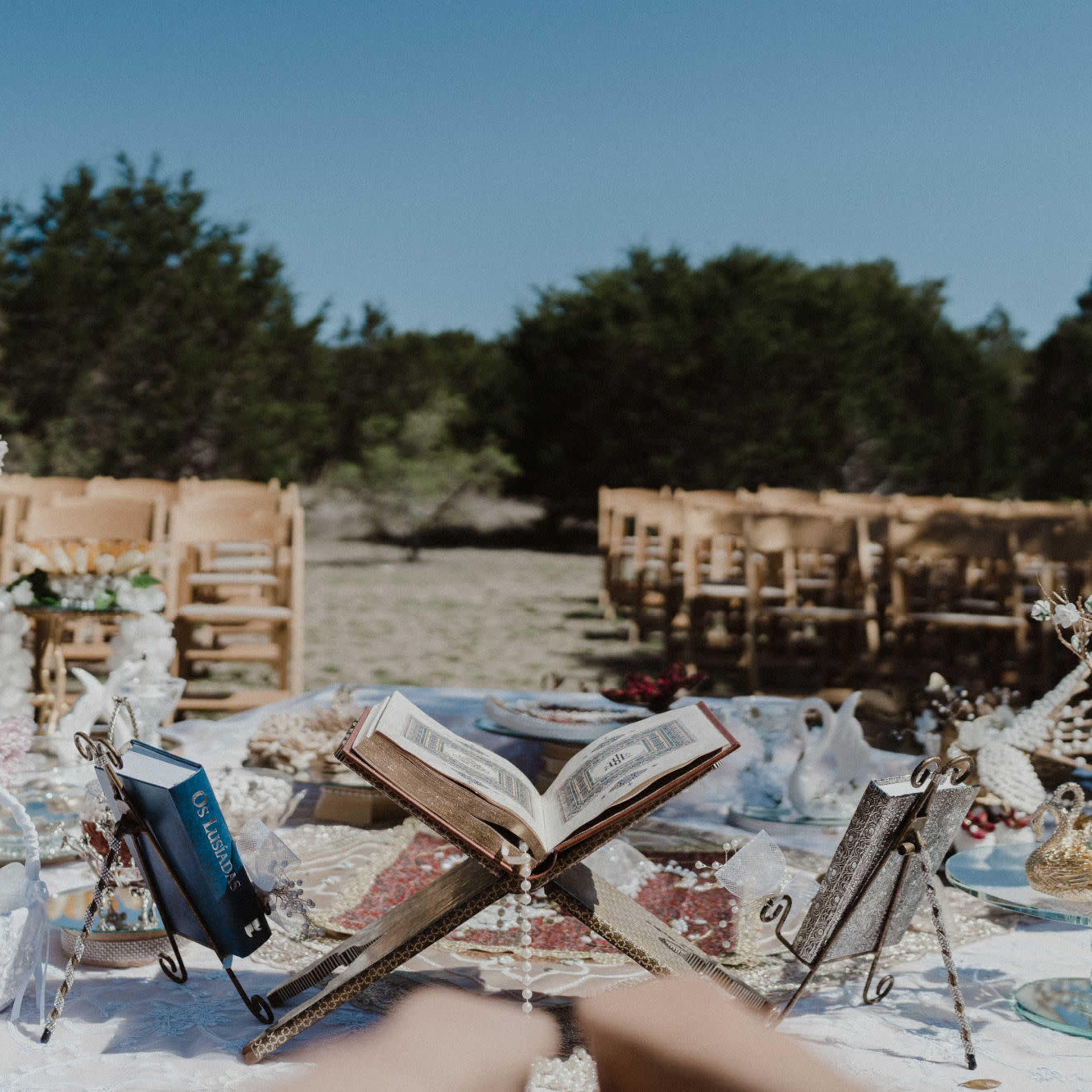 A wedding setup features decorated tables with an open book and various decorative items, with wooden chairs arranged in the background under a clear sky.