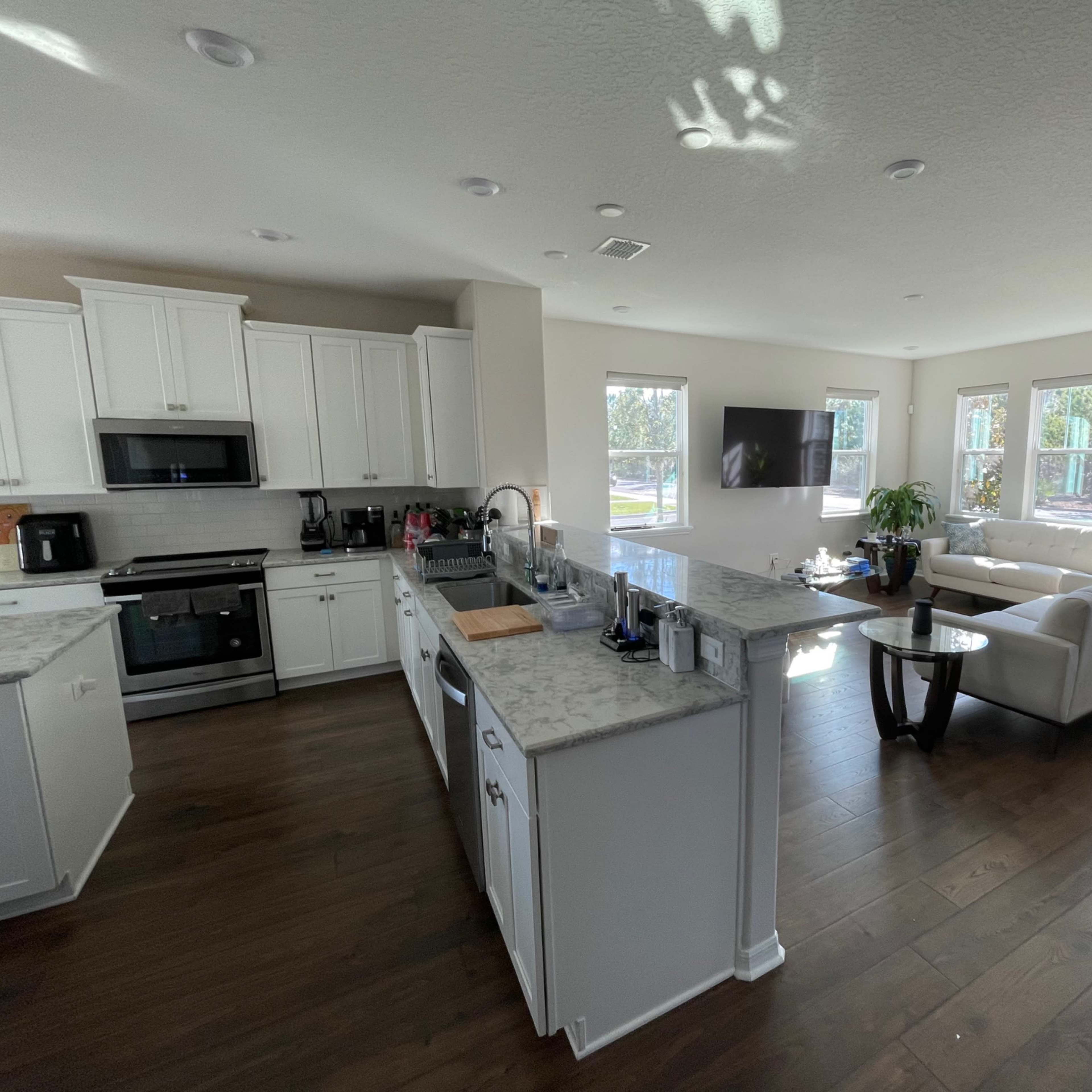 A modern open-concept living space features a kitchen with white cabinets and a marble countertop adjacent to a seating area with a sofa and large windows.