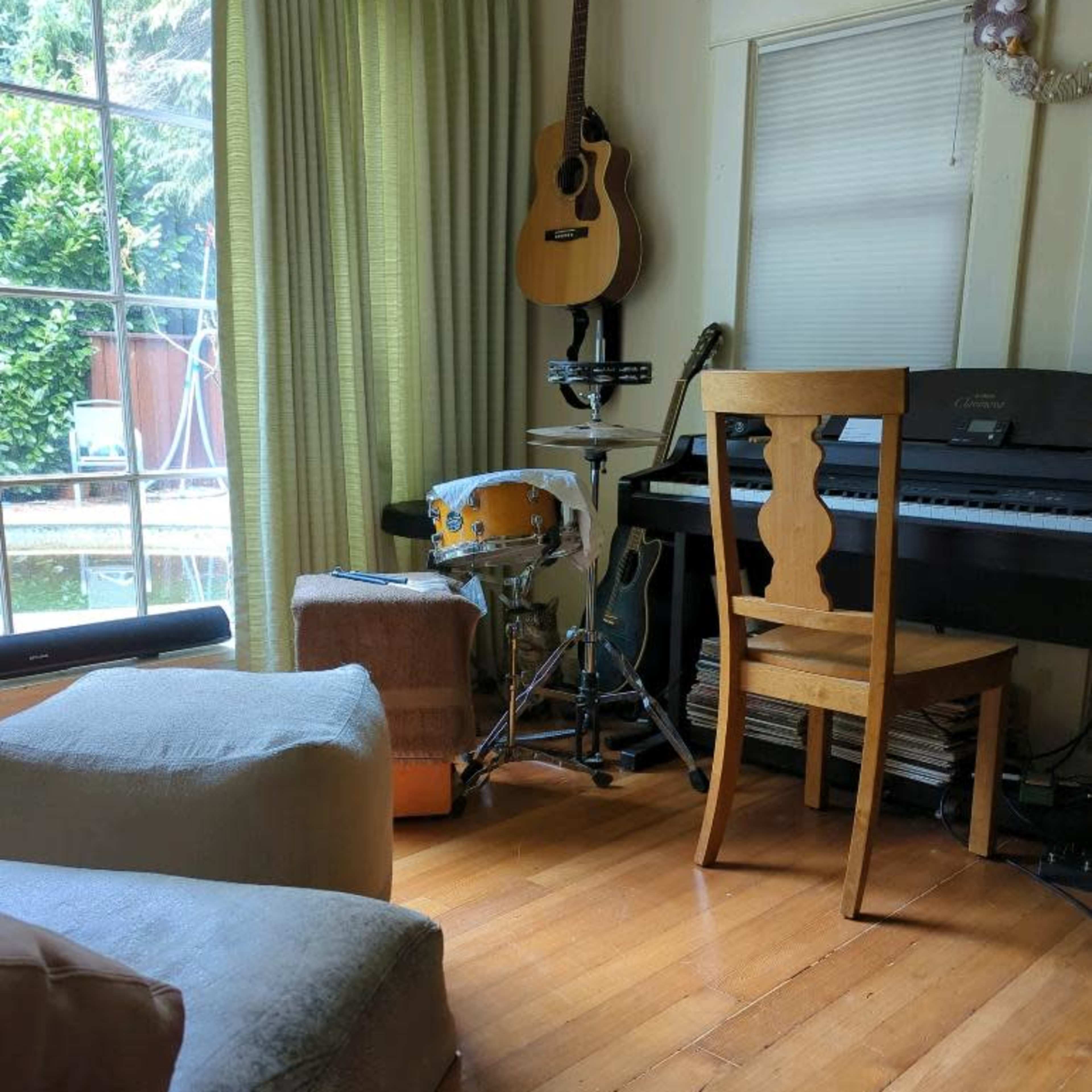A living room features a chair near a keyboard and a drum set, with a guitar hanging on the wall and large windows letting in natural light.