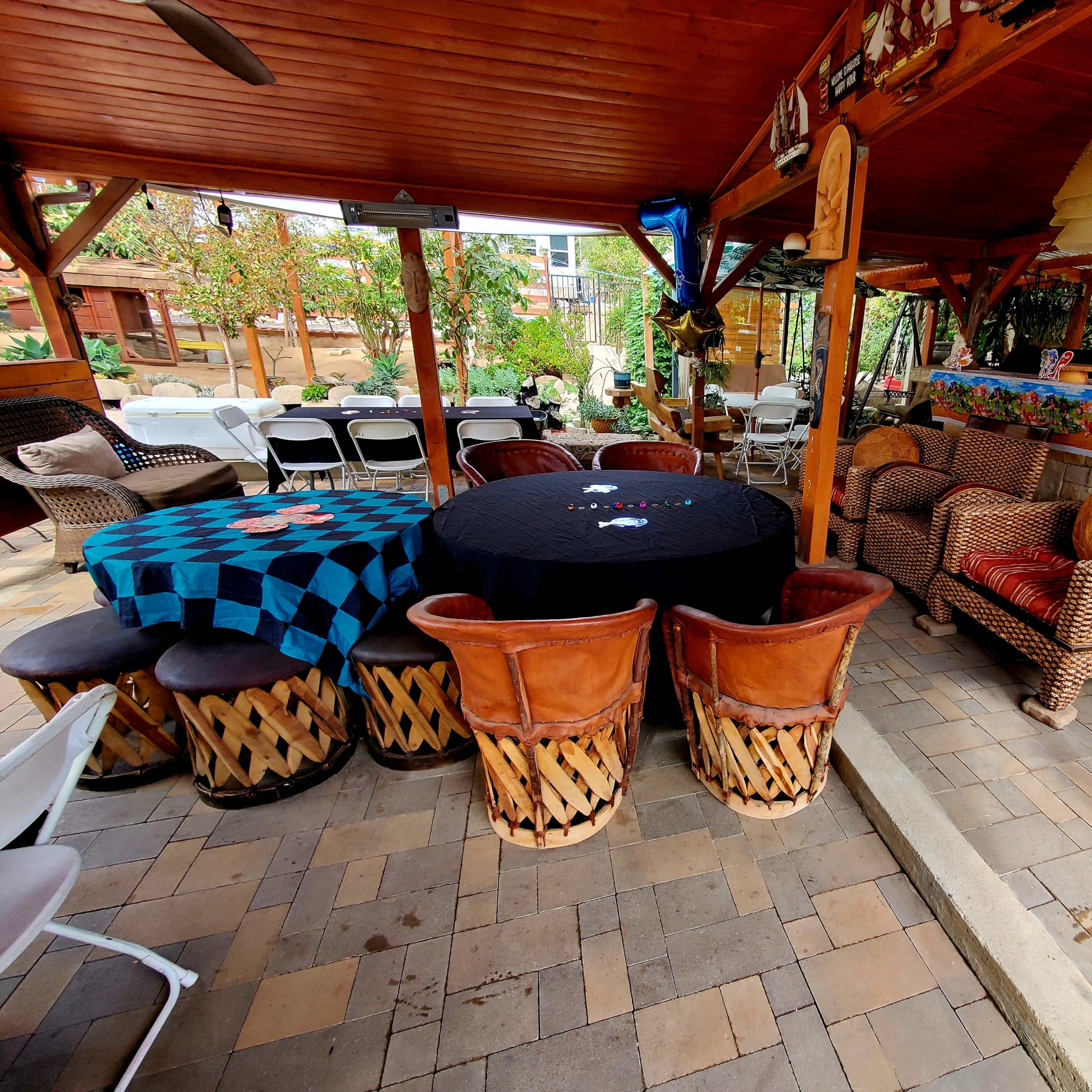 A covered outdoor seating area features a round table with a black tablecloth, surrounded by woven chairs and drum-style stools.
