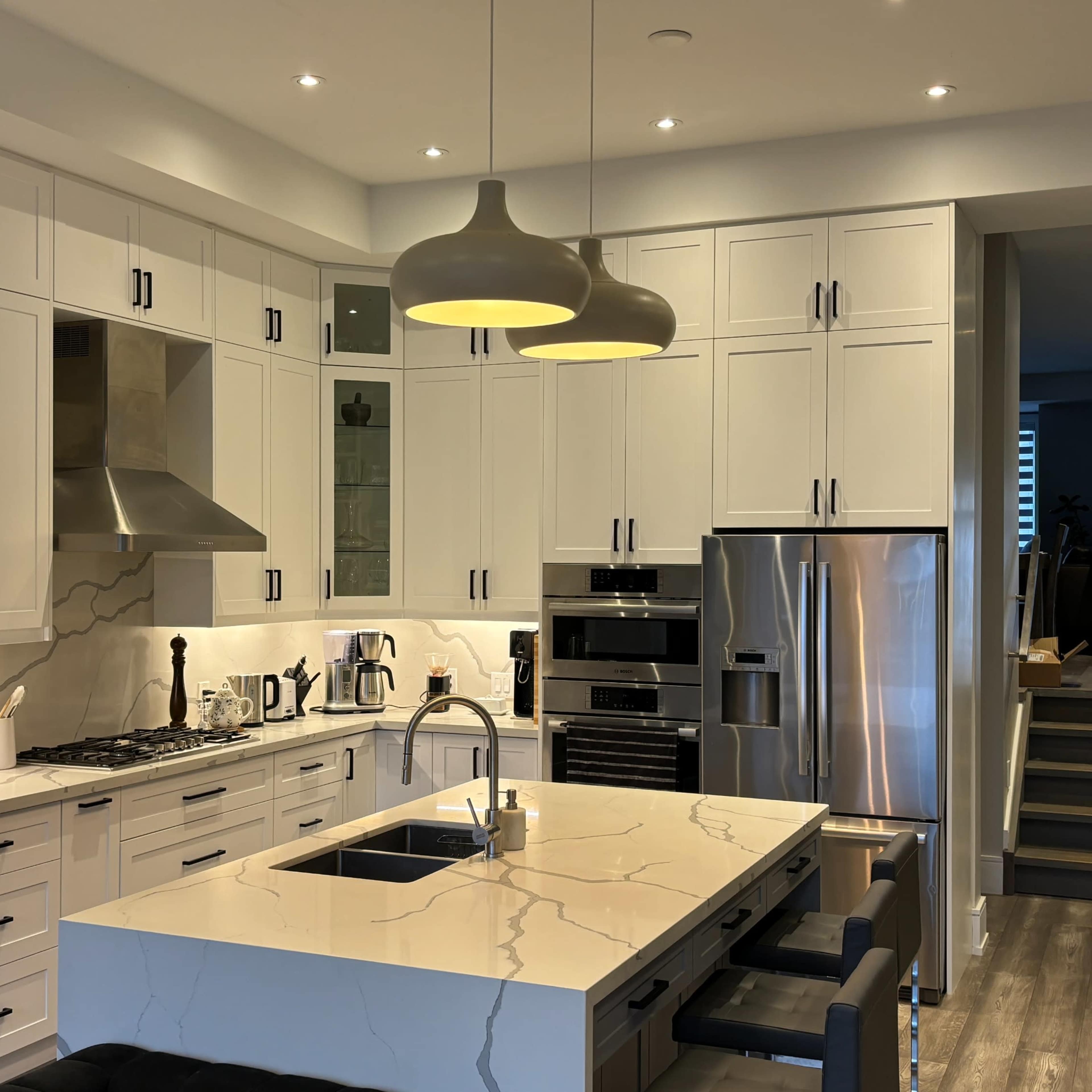 A modern kitchen features white cabinetry, a large island with a marble countertop, and pendant lighting above, leading to a staircase in the background.