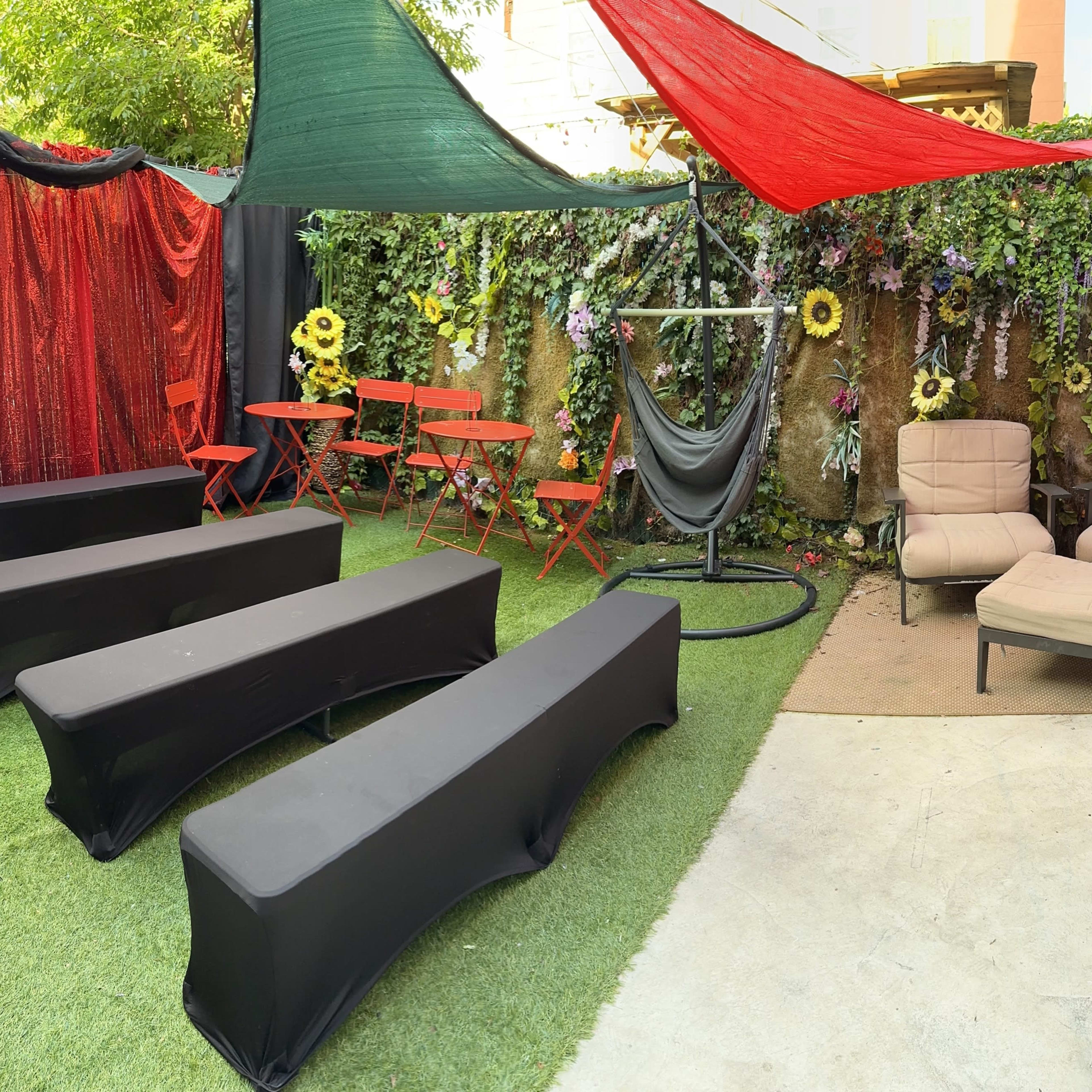 A colorful outdoor space featuring black seating, red and green canopies, and floral decorations, with two armchairs and a hammock in a corner.