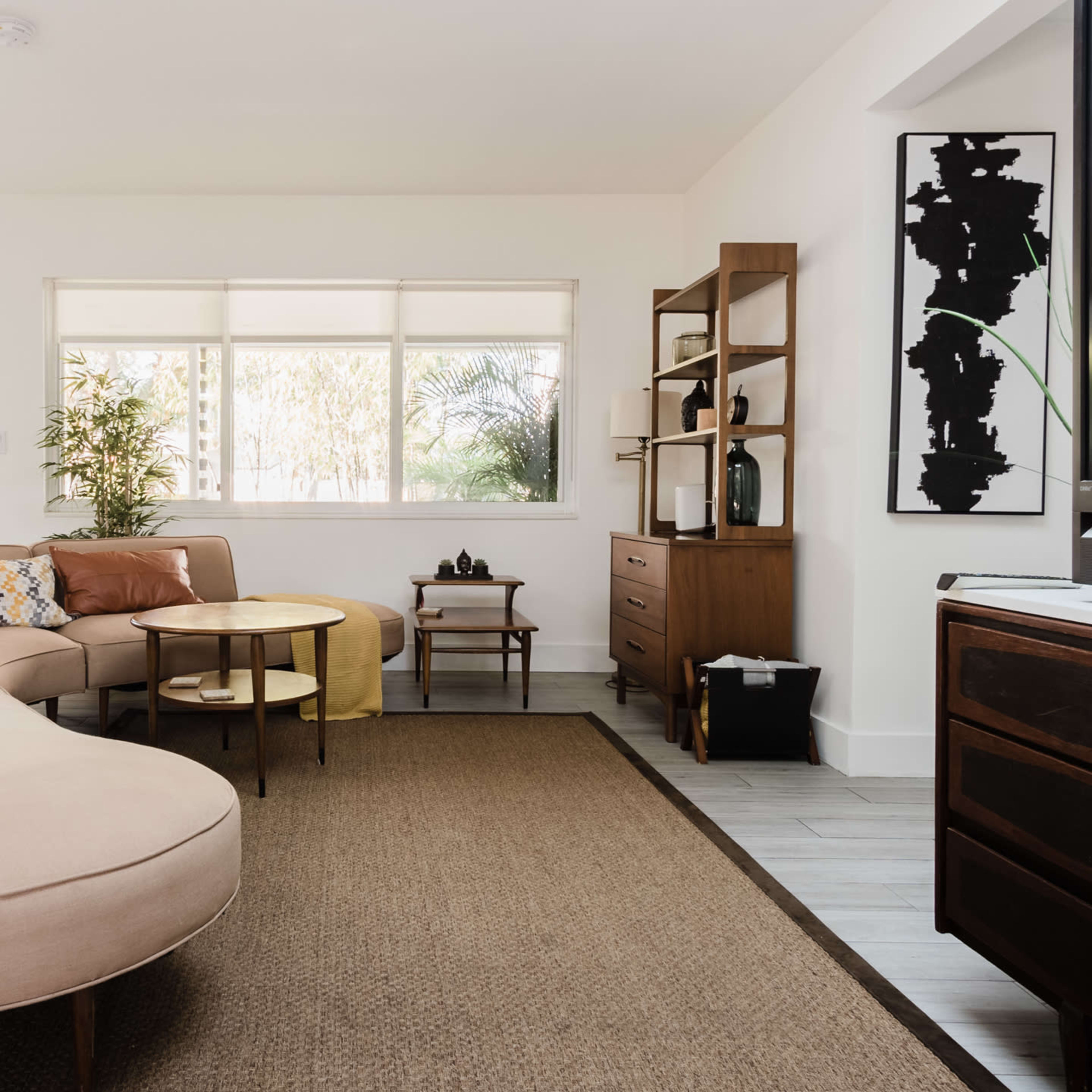 A modern living room featuring a curved beige sofa, two coffee tables, and a wooden shelving unit against light-colored walls and large windows.