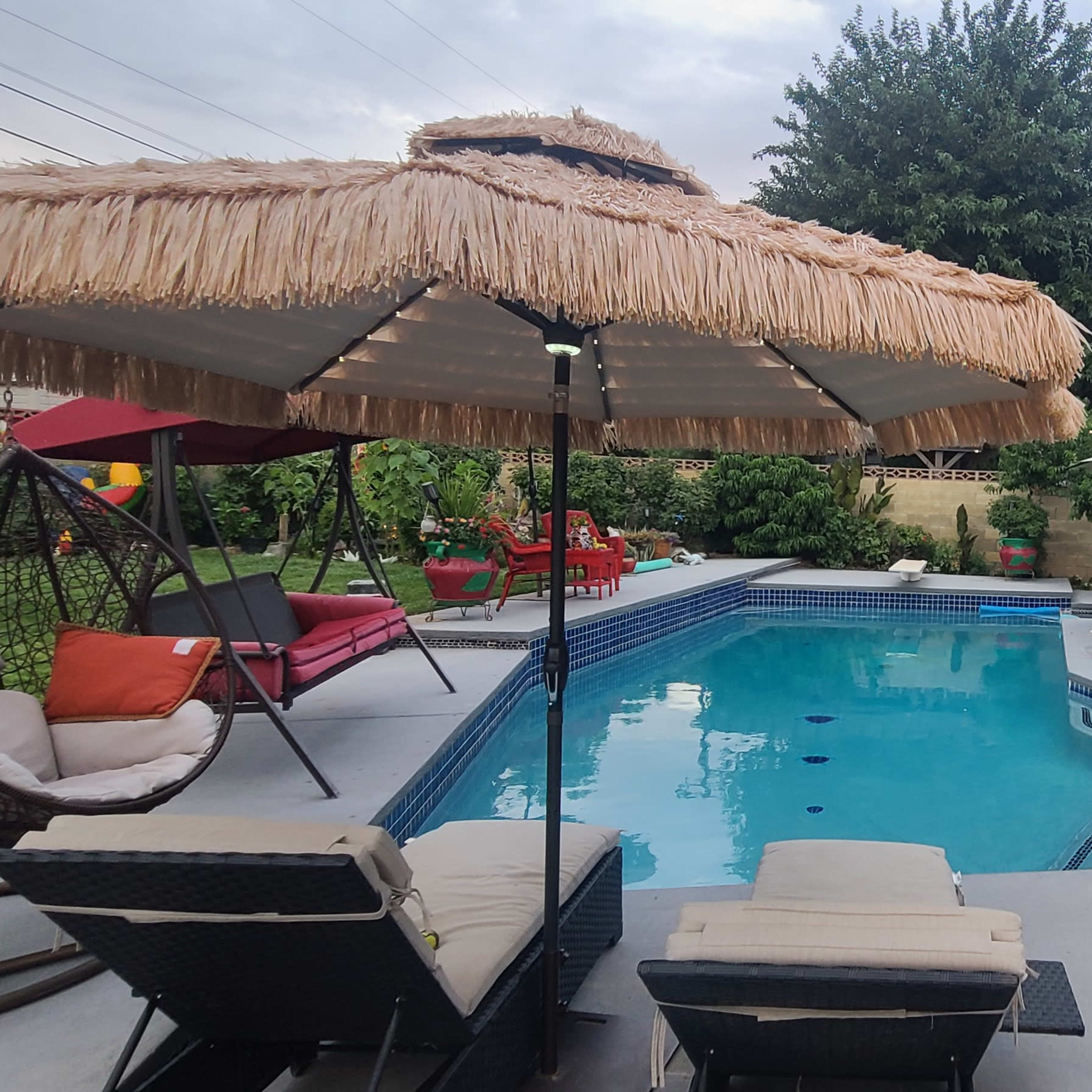 A backyard pool area featuring lounge chairs, umbrellas, and various seating arrangements surrounded by greenery and a house.