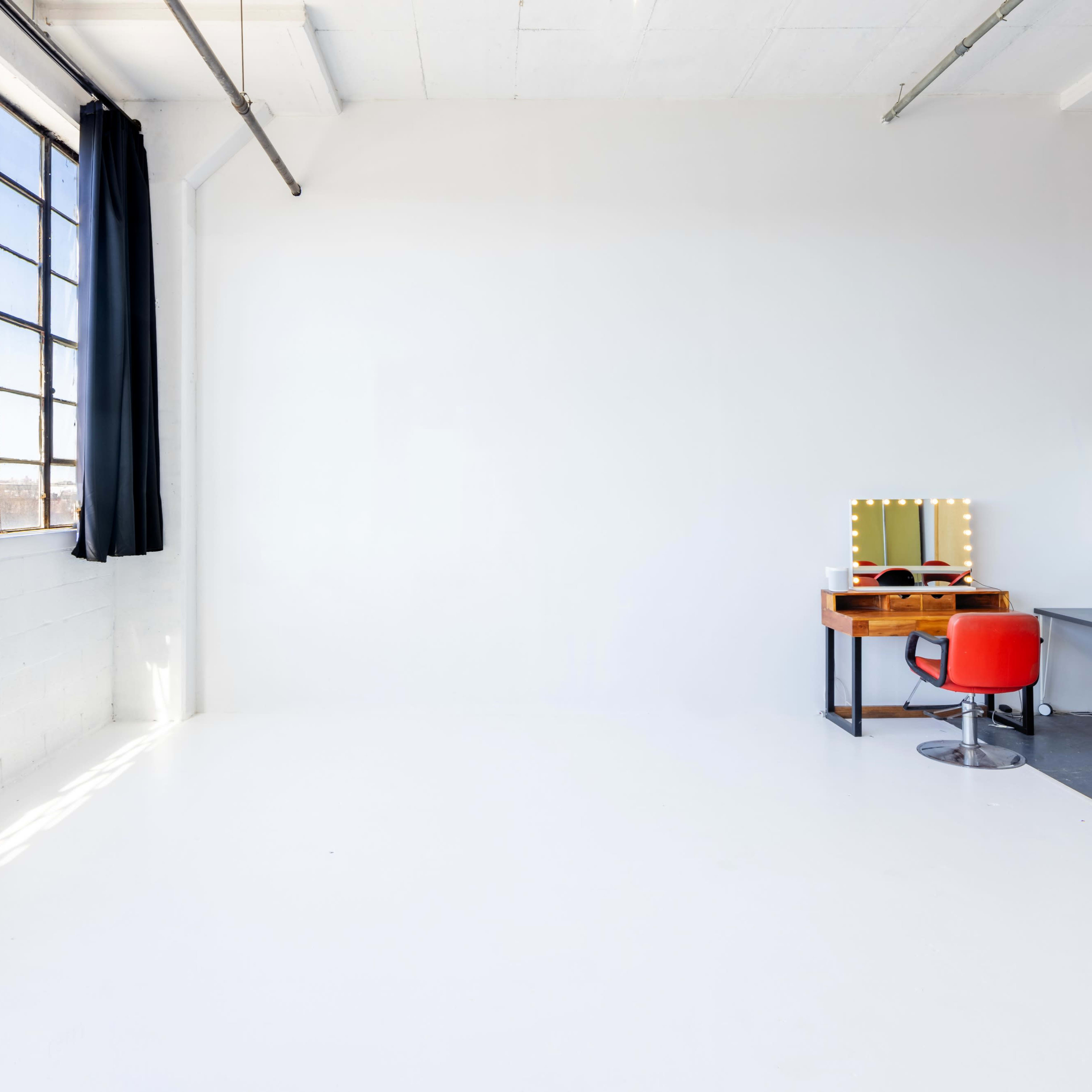 A bright, empty studio space features large windows, a wall with white paint, and a dressing table with a red chair.