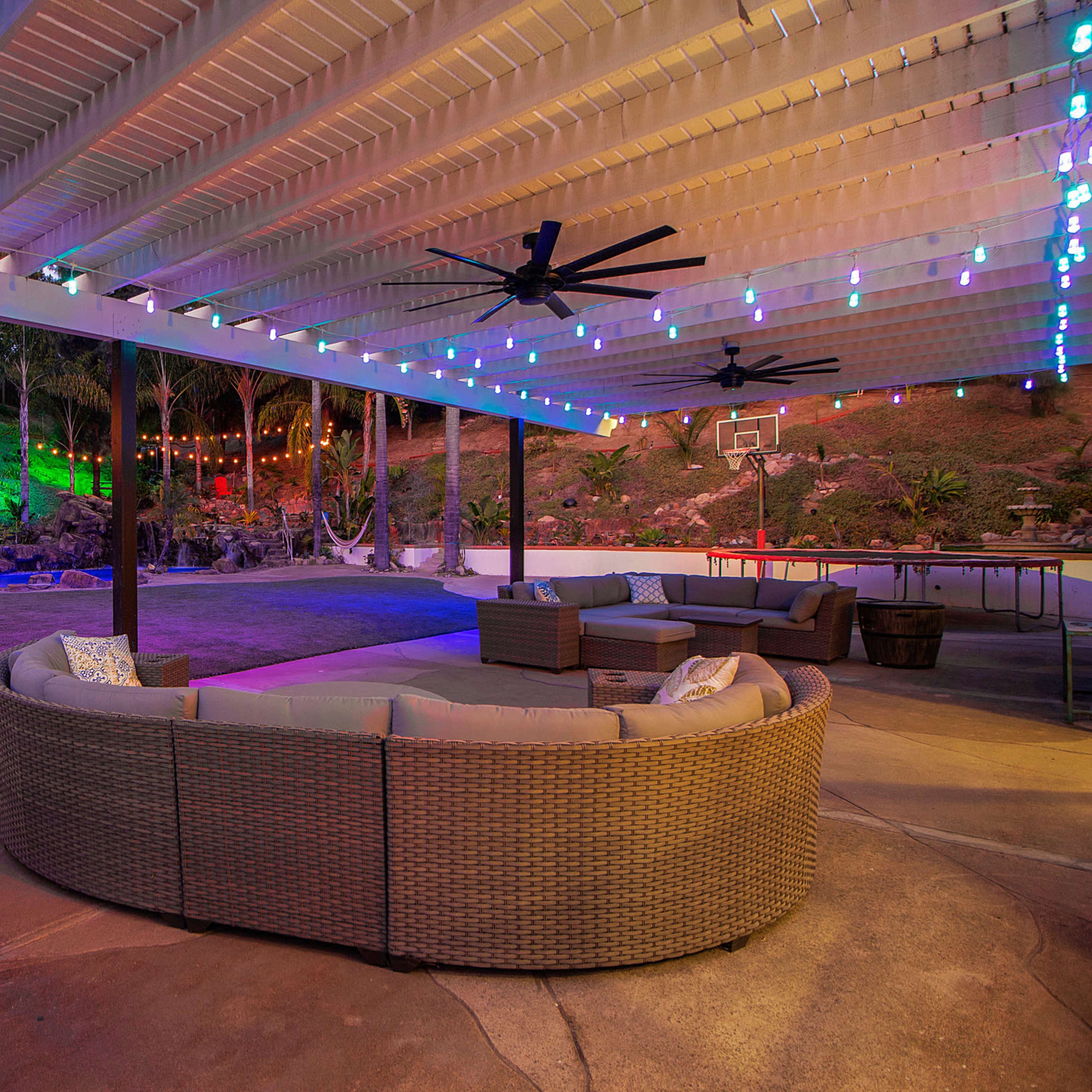 A spacious outdoor lounge area with a circular sectional sofa under a covered patio, featuring colorful string lights and a basketball hoop in the background.