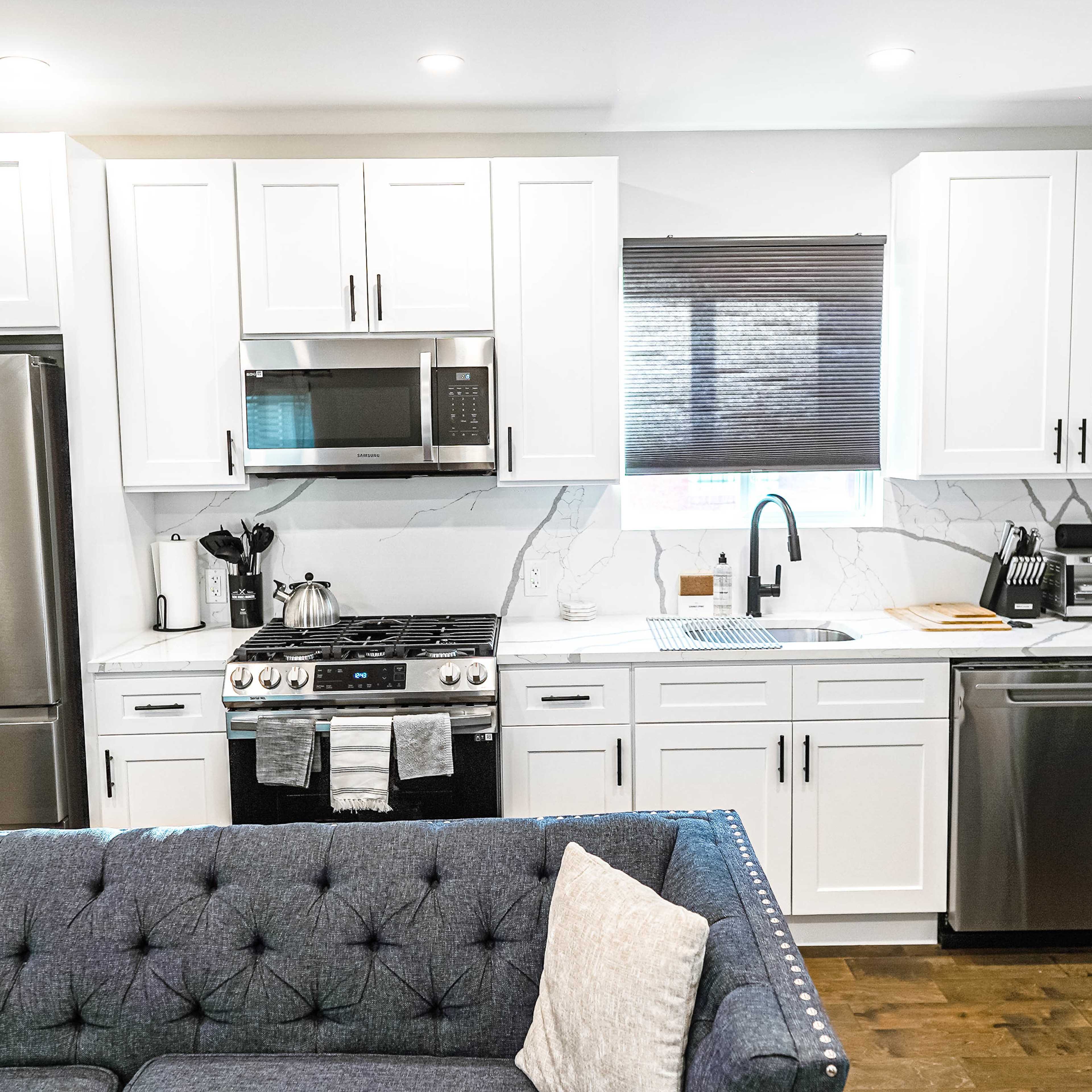 A modern kitchen with white cabinets, stainless steel appliances, and a marble countertop, adjacent to a blue tufted sofa.