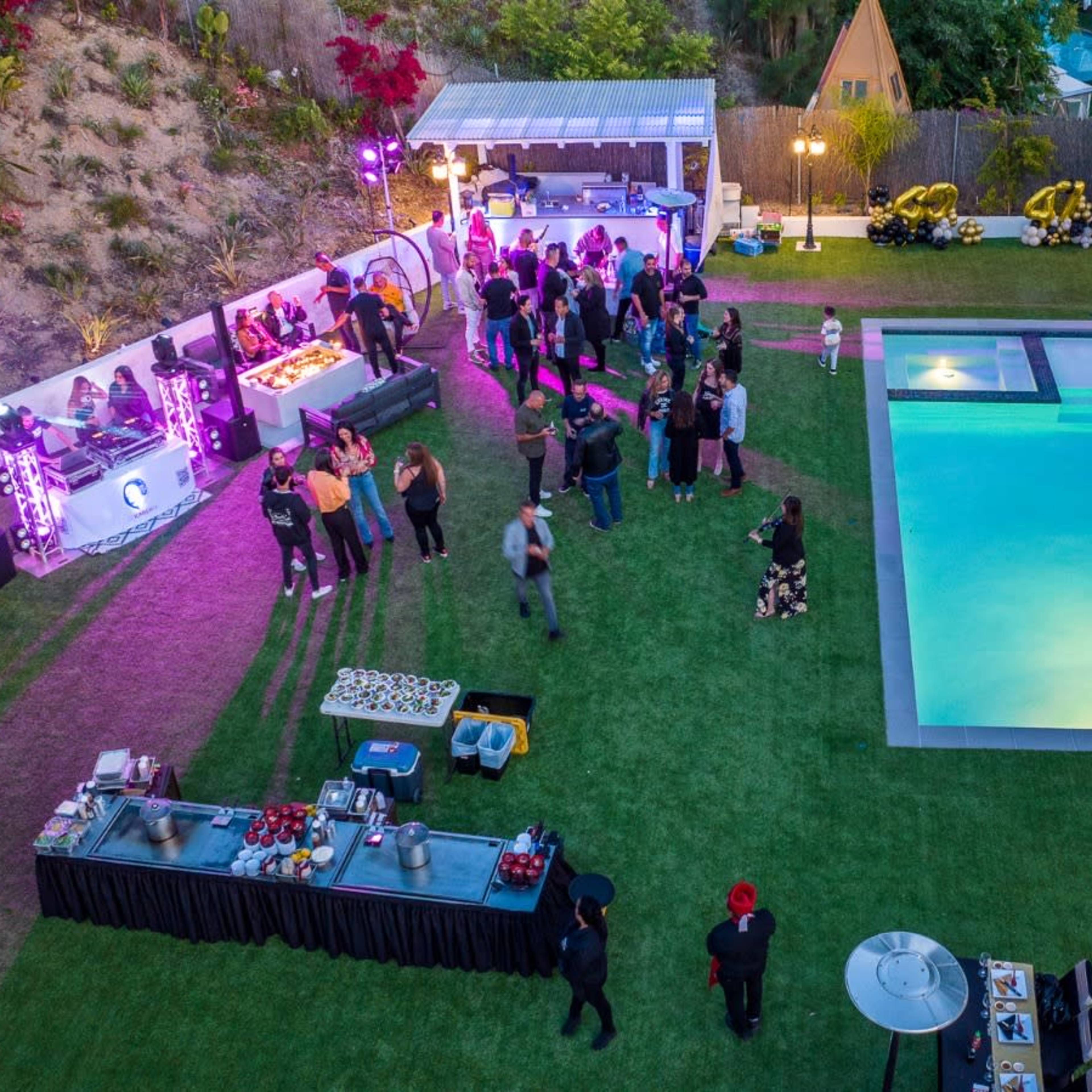 A large outdoor gathering takes place around a swimming pool, with guests mingling near food and drink stations in a landscaped backyard.