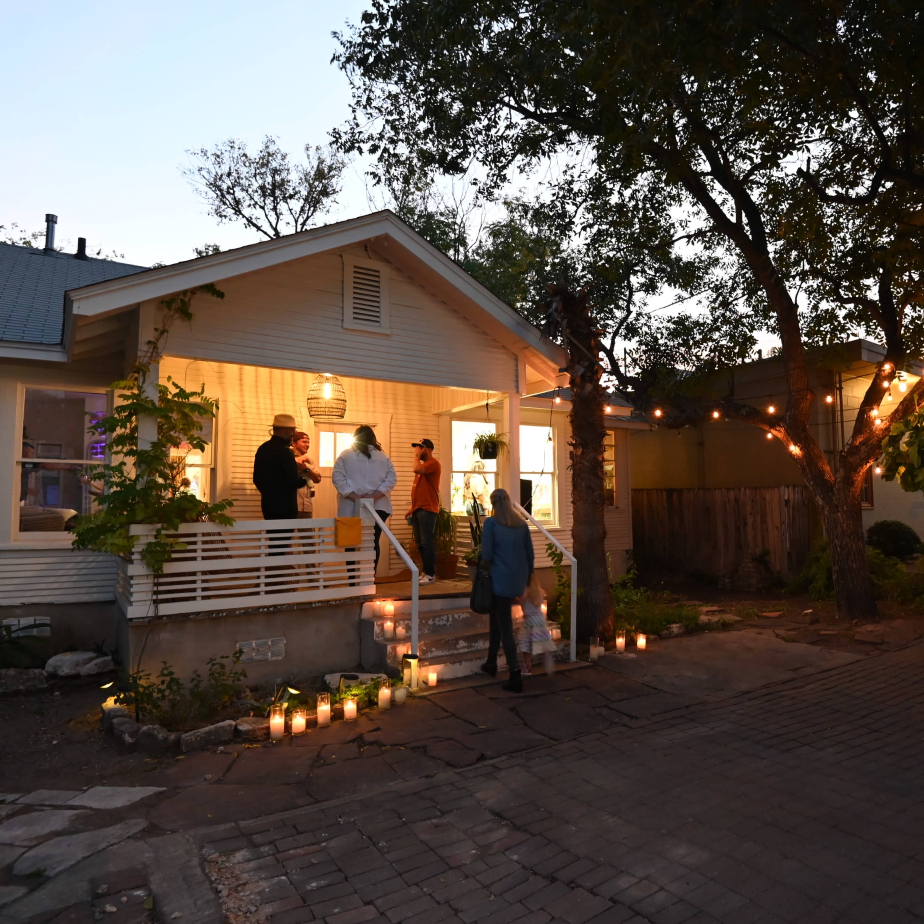 A group of people gathers on the porch of a well-lit house with candles lining the pathway as twilight approaches.