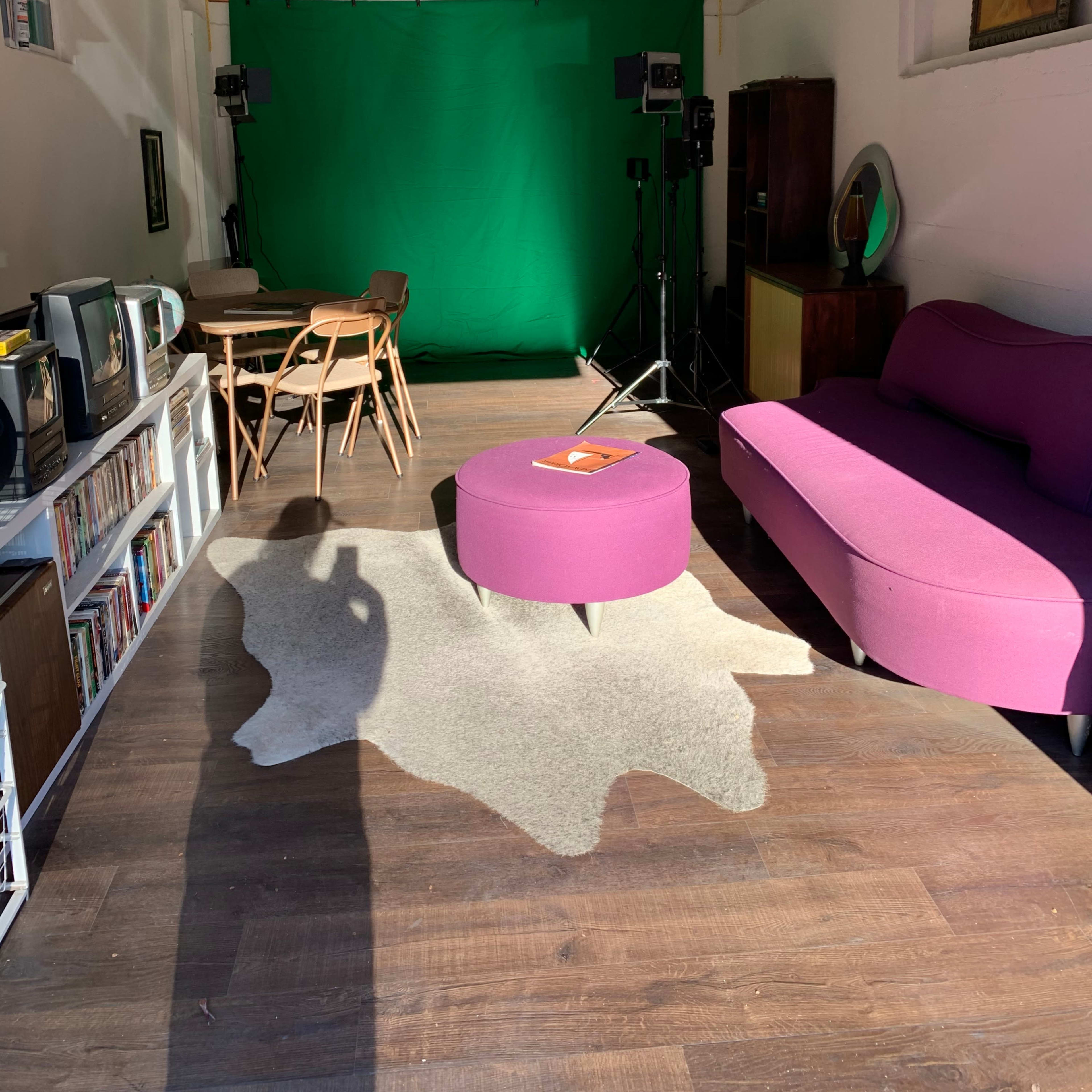 A modern room features a green backdrop with studio lights, a pink sofa, a circular purple coffee table, and a wooden dining set.