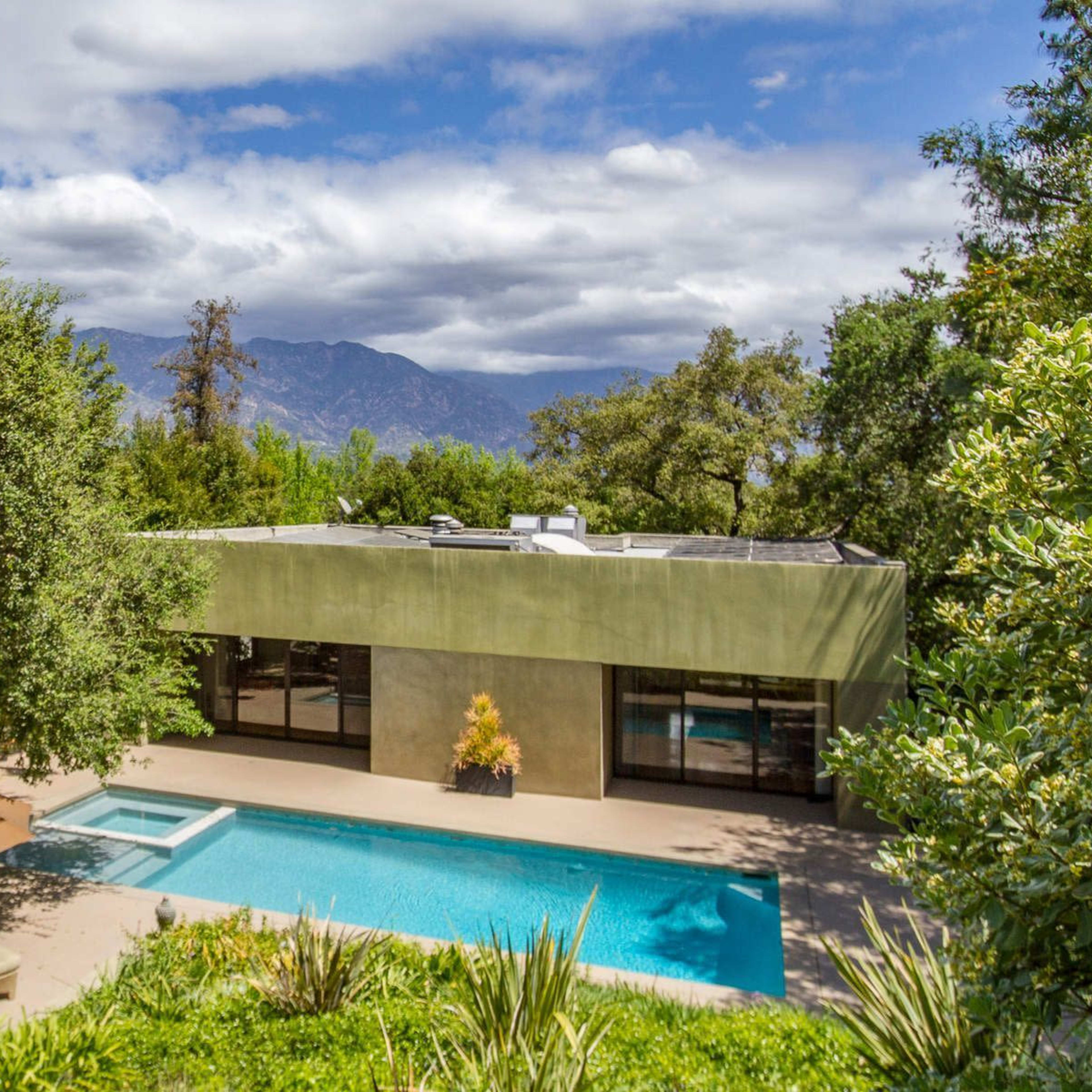 A modern house with large windows overlooks a swimming pool surrounded by greenery and mountains in the background.