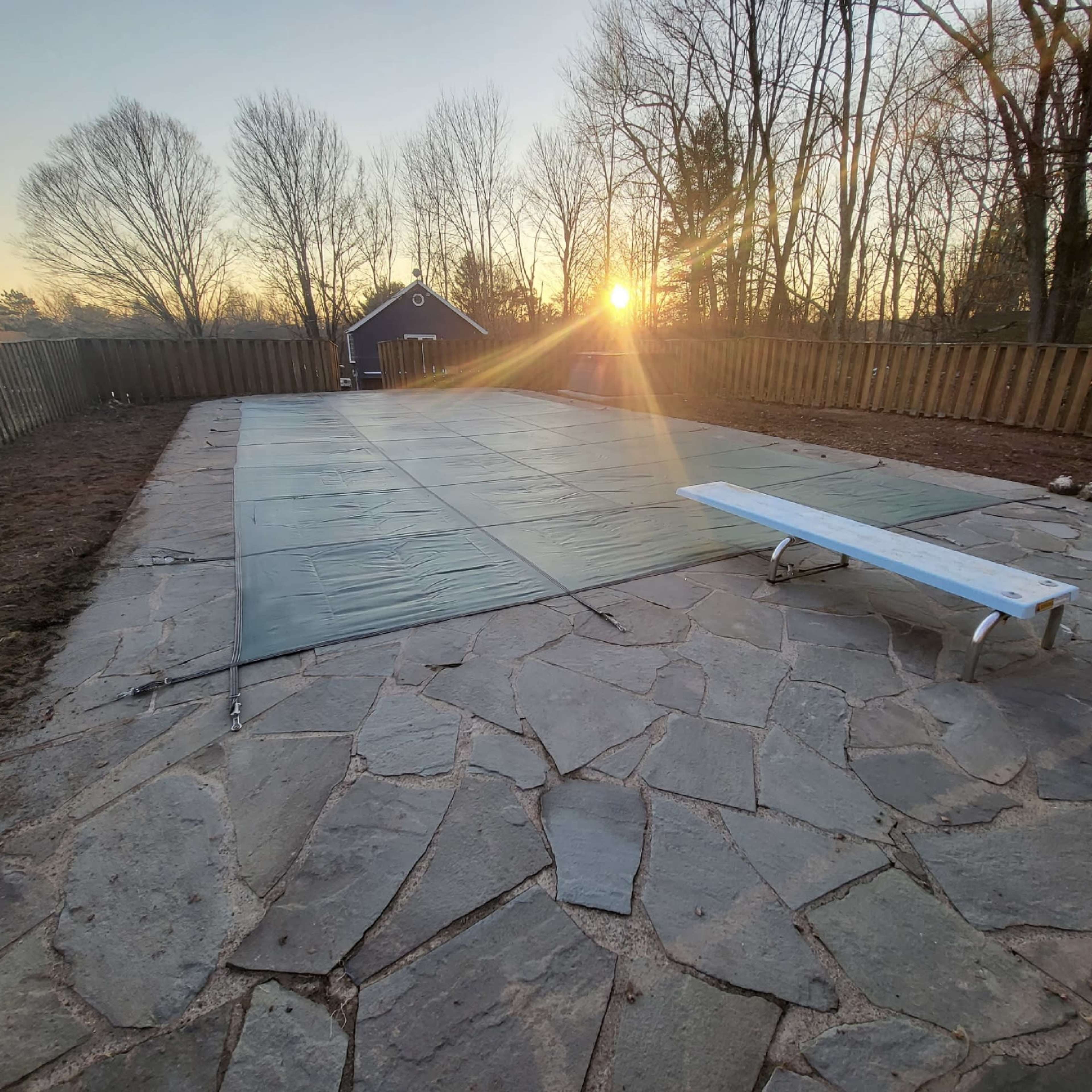 A pool area covered with a green safety cover, surrounded by a stone patio and a wooden fence, with a setting sun in the background.